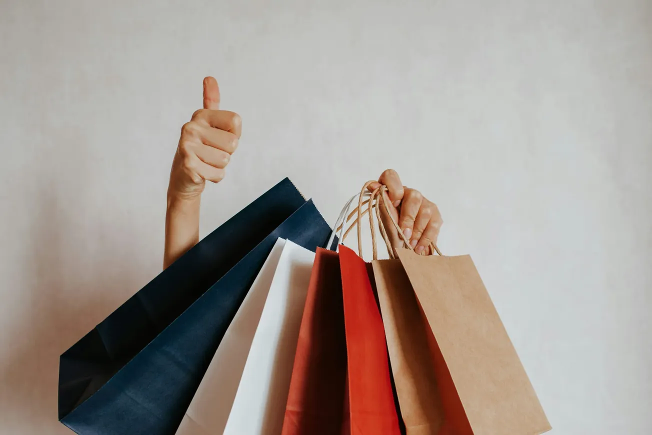 A hand holds colorful shopping bags in red, blue, white, and brown, with another hand showing a thumbs-up sign, conveying satisfaction and success.