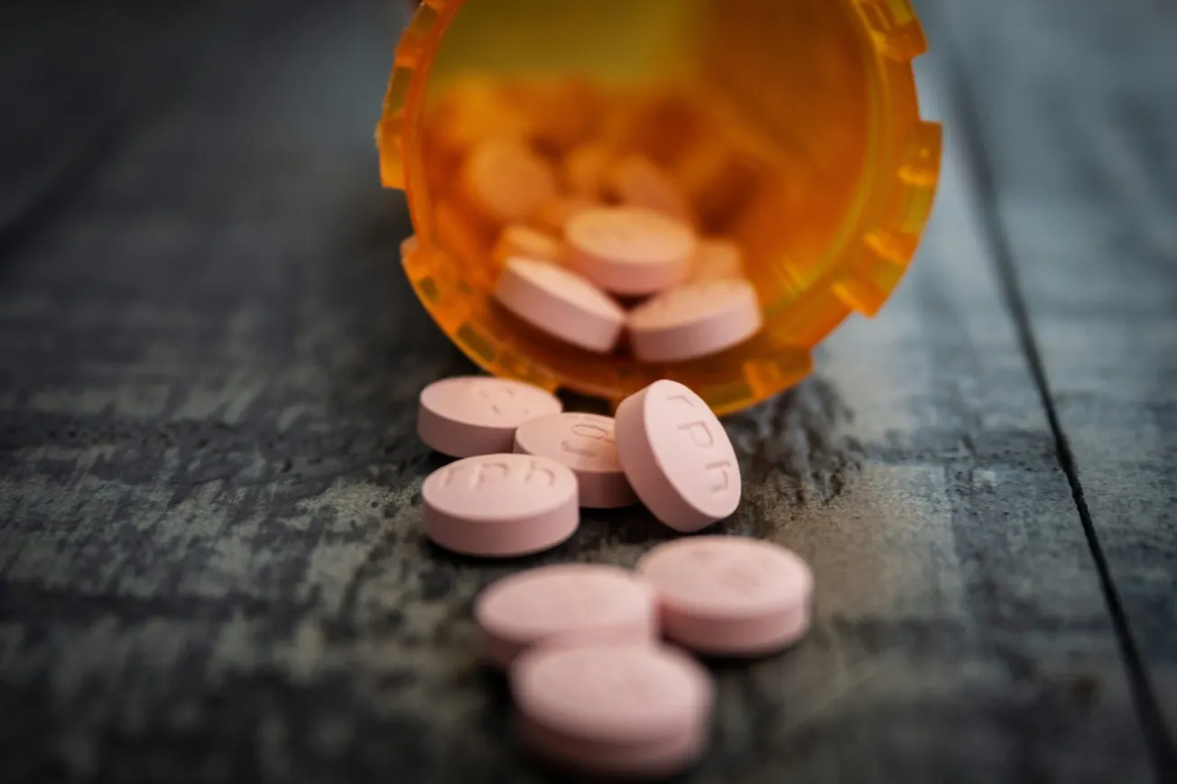 A tipped-over orange pill bottle spills pink tablets onto a wooden surface. The scene conveys medication or health-related themes, hinting at care or caution.