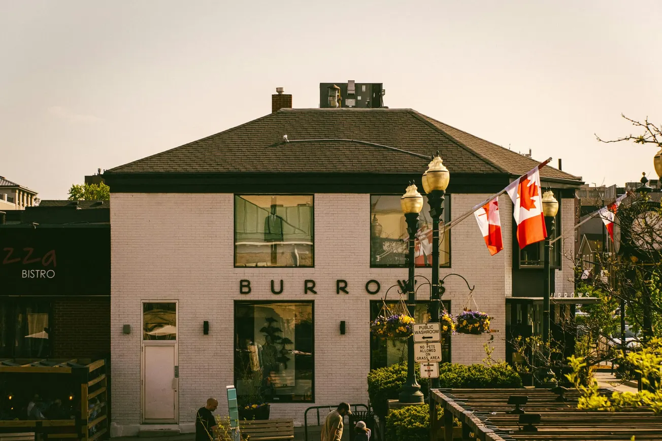 A two-story building labeled "BURRO" with large windows is adorned with Canadian flags. The scene is vibrant and lively in a sunny urban setting.