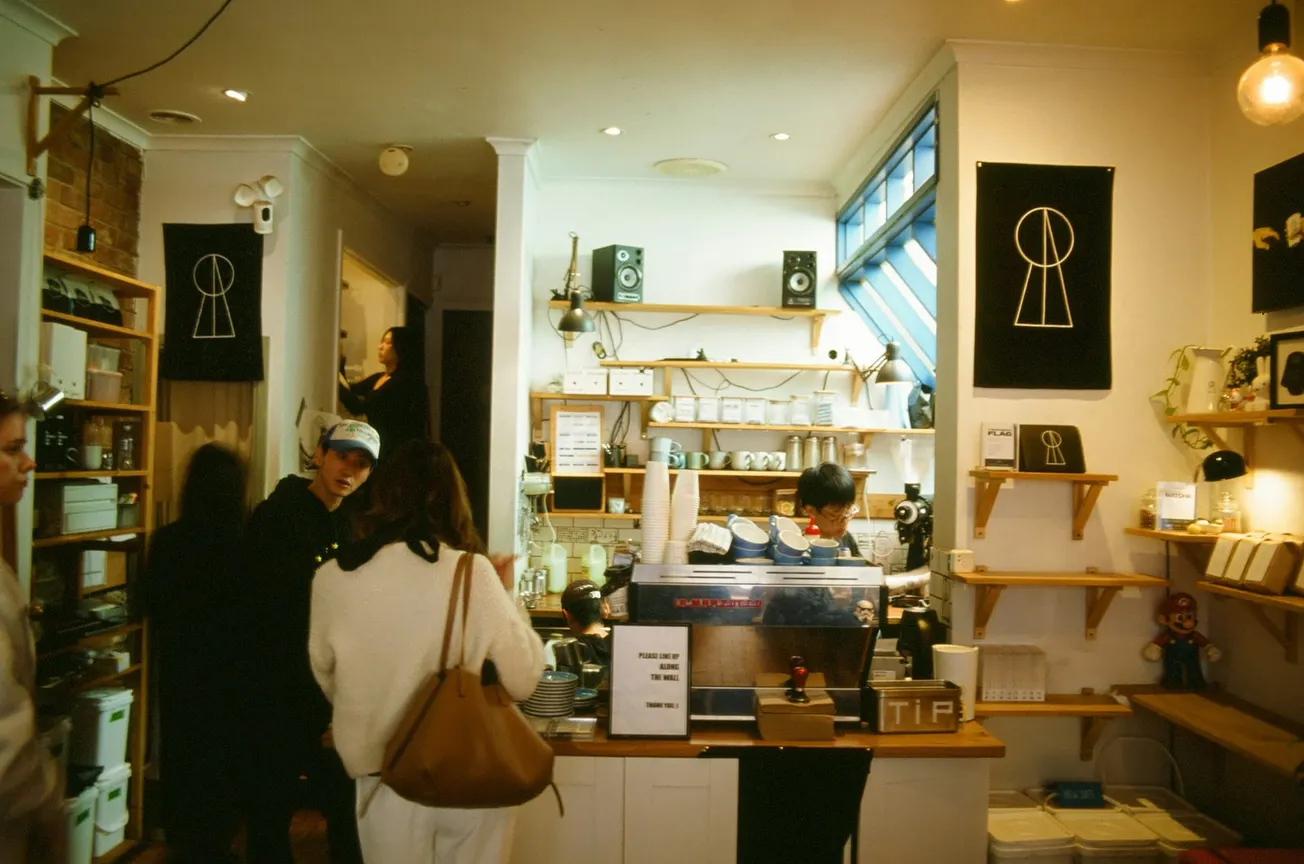Cozy café interior with warm lighting. A barista works behind an espresso machine, while patrons browse. Shelves hold mugs and décor, creating an inviting atmosphere.