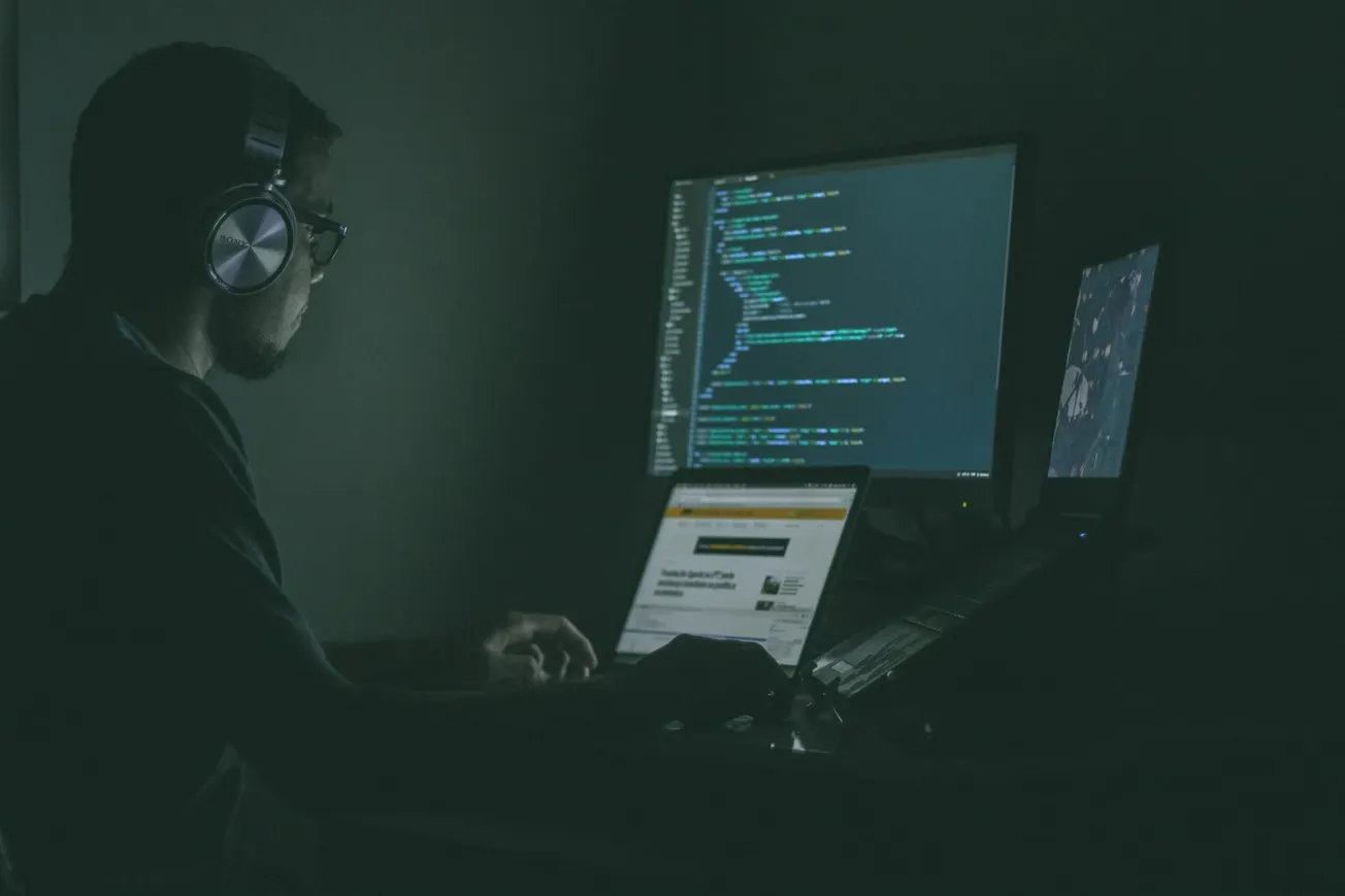 A person wearing headphones works in a dimly lit room with glowing computer screens displaying code, conveying focus and concentration.