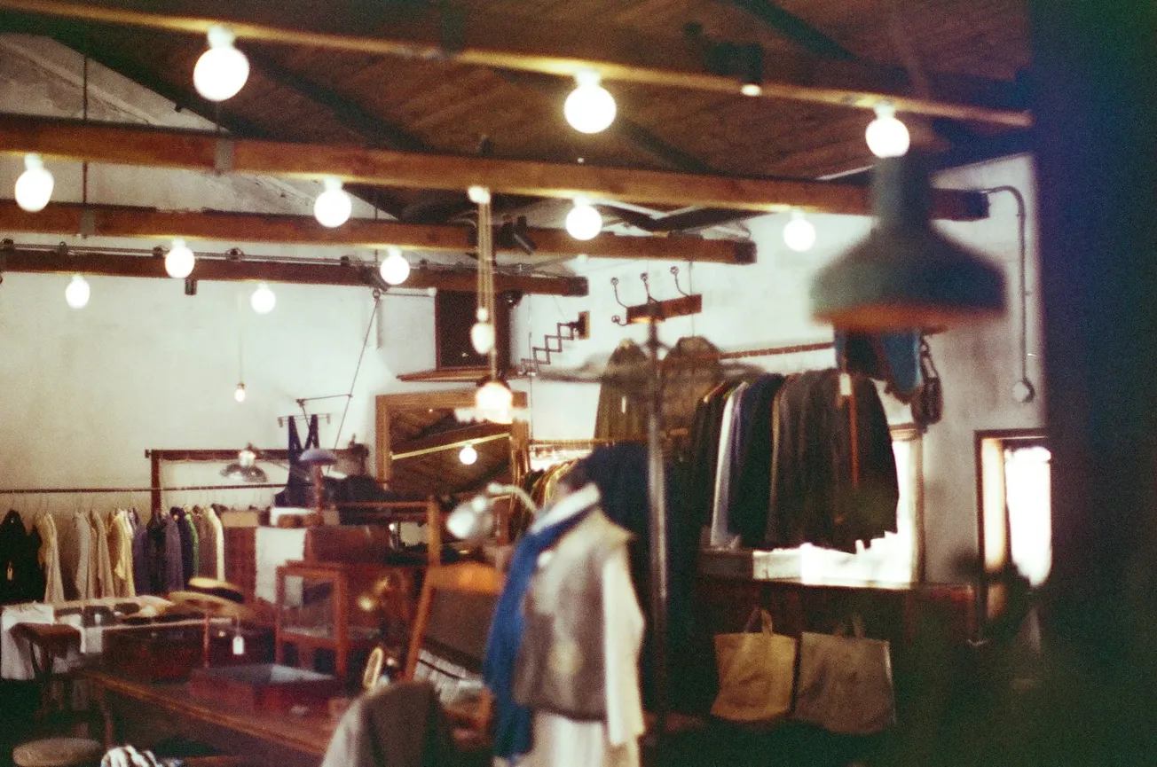 Dimly lit vintage clothing store with wooden beams and hanging bulbs. Racks display various apparel, and bags are placed on a table, creating a nostalgic atmosphere.