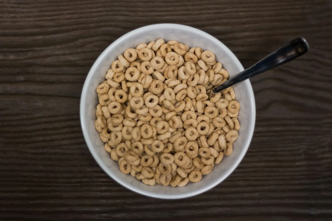 A white bowl filled with dry, ring-shaped cereal sits on a dark wooden surface. A silver spoon rests inside, suggesting breakfast.