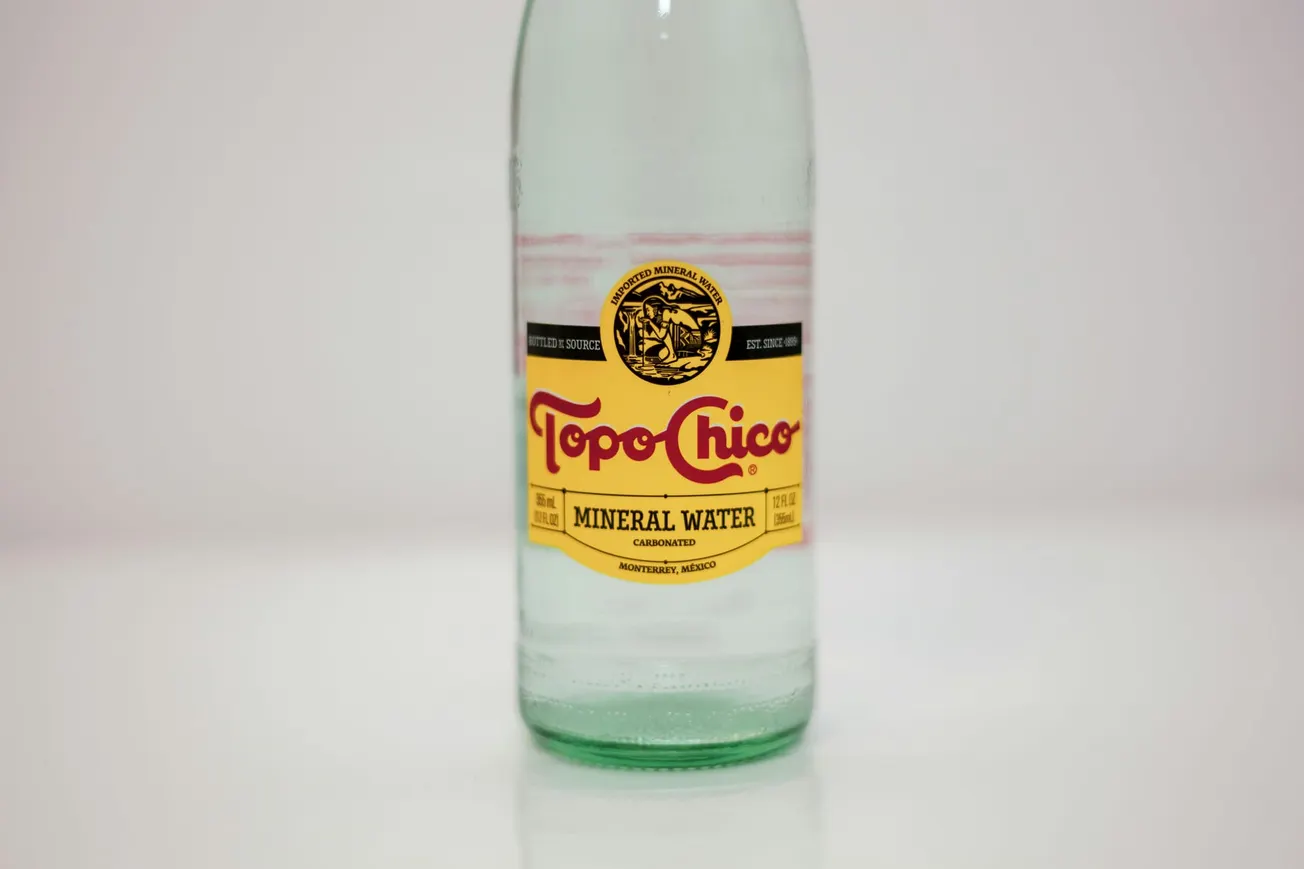 Clear glass bottle of Topo Chico mineral water on a white background. The label is yellow with red text and a black emblem, conveying a classic, refreshing tone.