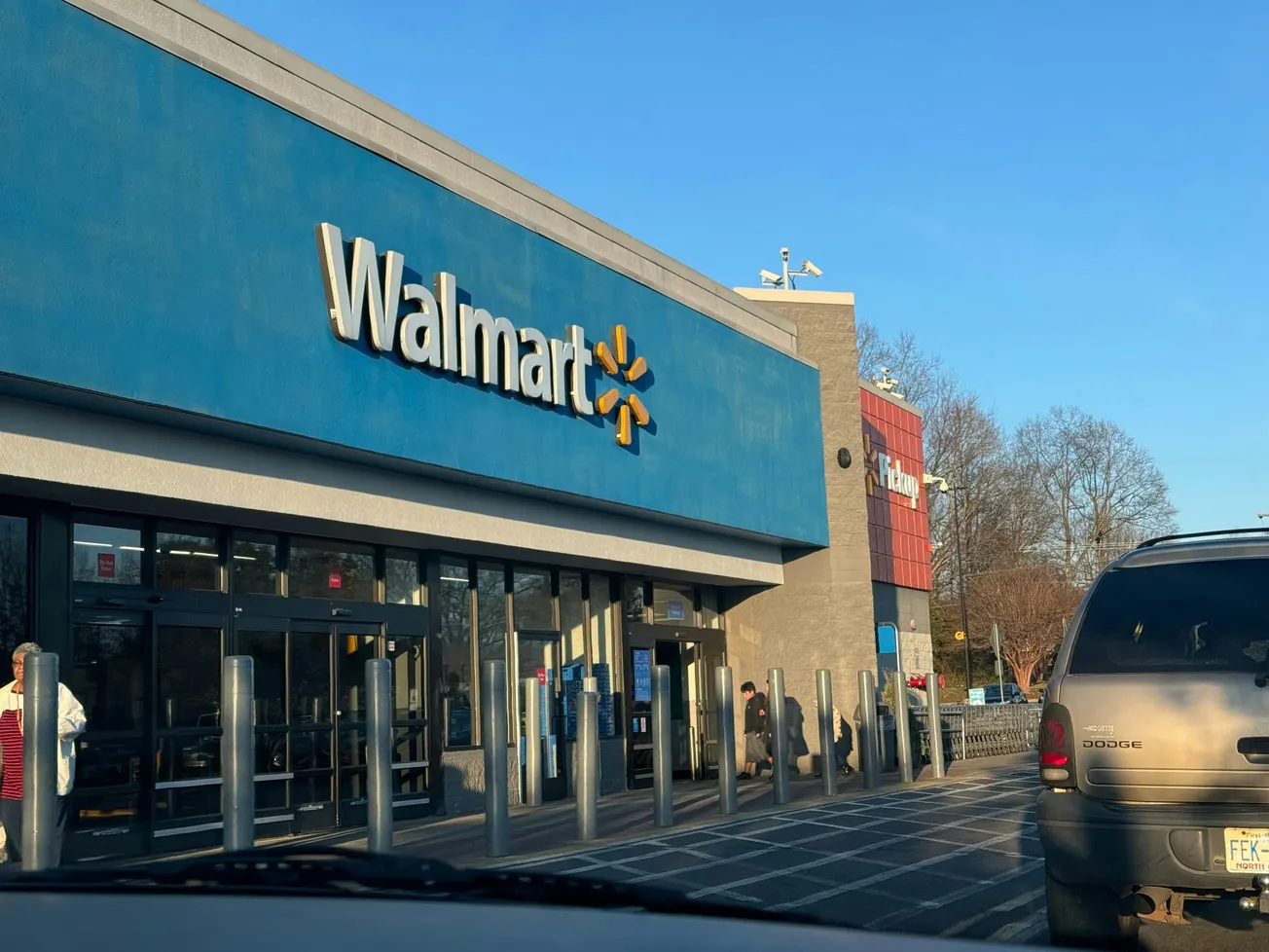 Walmart store entrance with a blue sign on a sunny day. People walk near the entrance, a gray SUV is driving in the foreground, trees in the background.