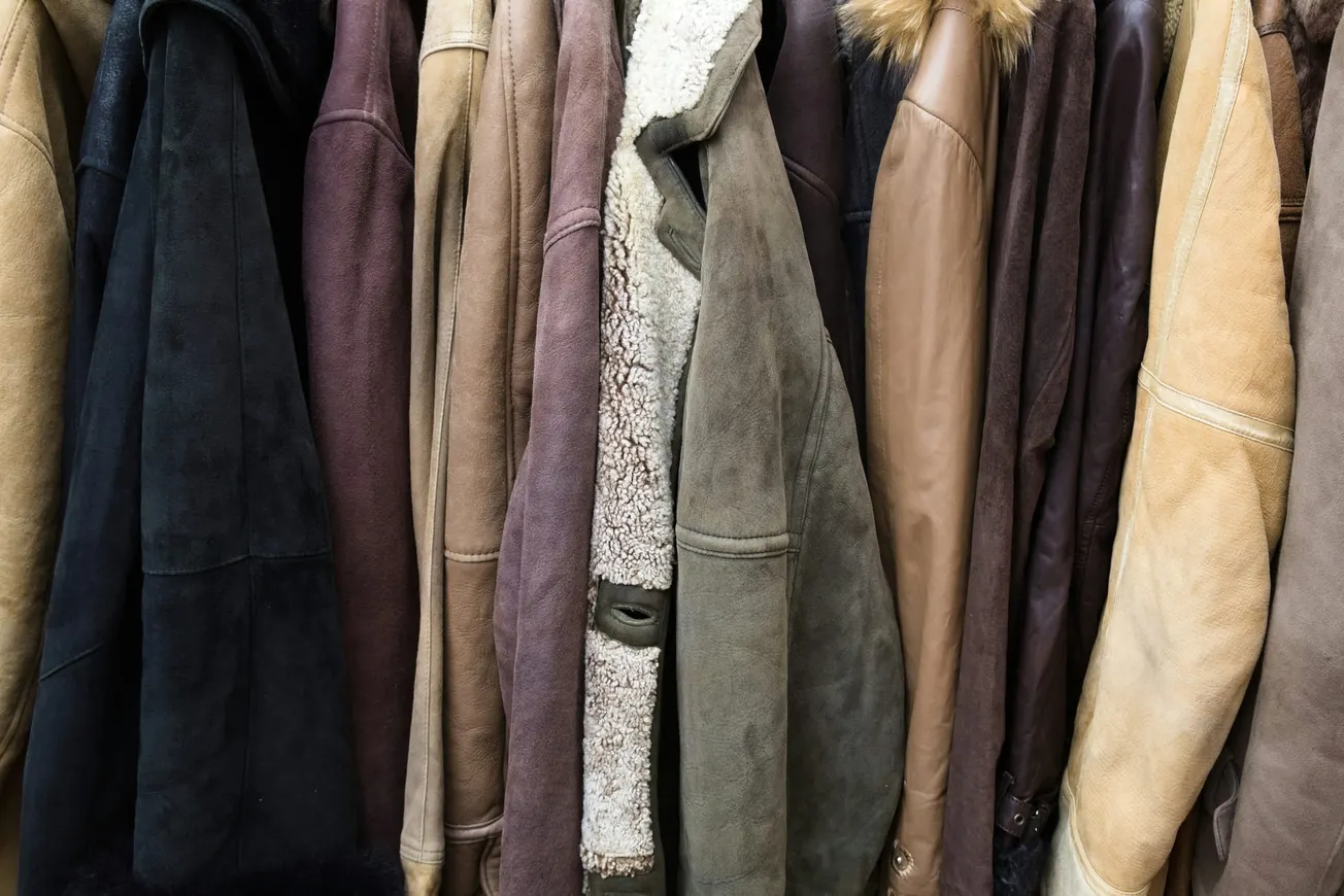 A row of various leather and suede jackets in earthy tones hangs closely together, showcasing textures like fur collars and shearling linings, evoking warmth.