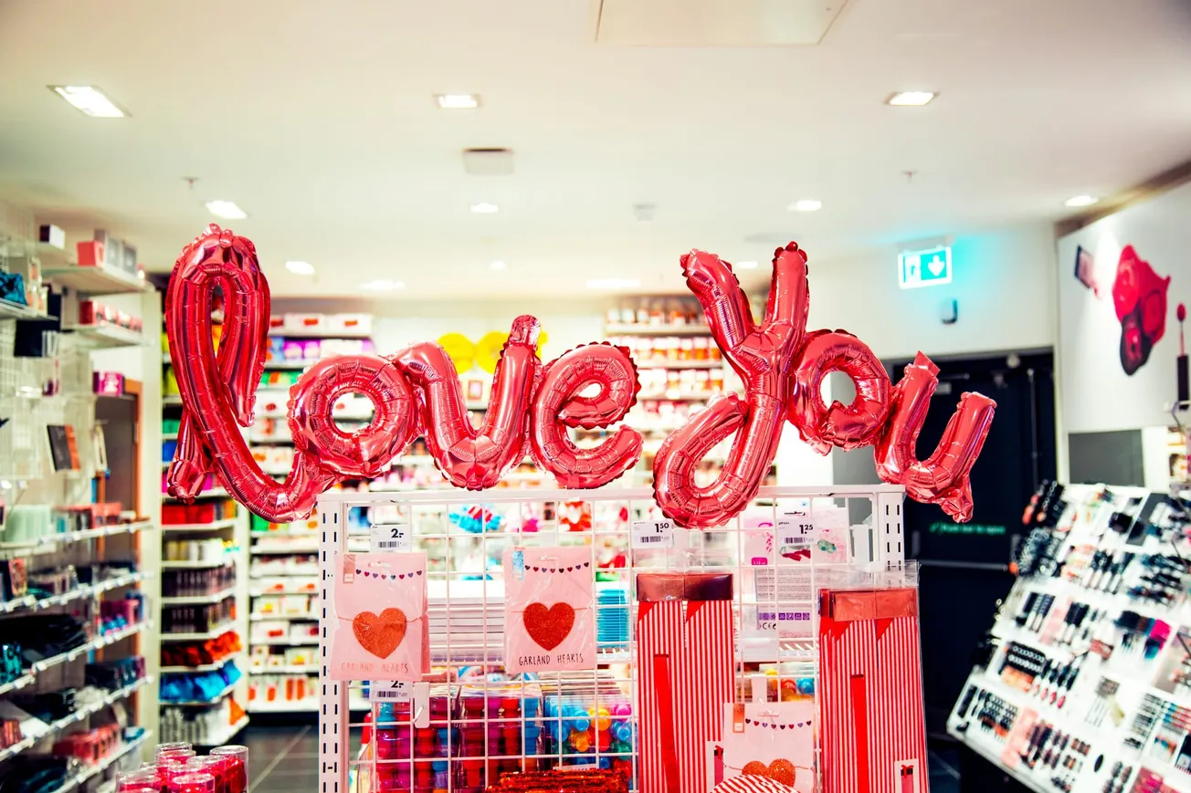 Red "love you" balloons in a bright store filled with colorful products. Gift bags with heart designs create a cheerful and romantic ambiance.