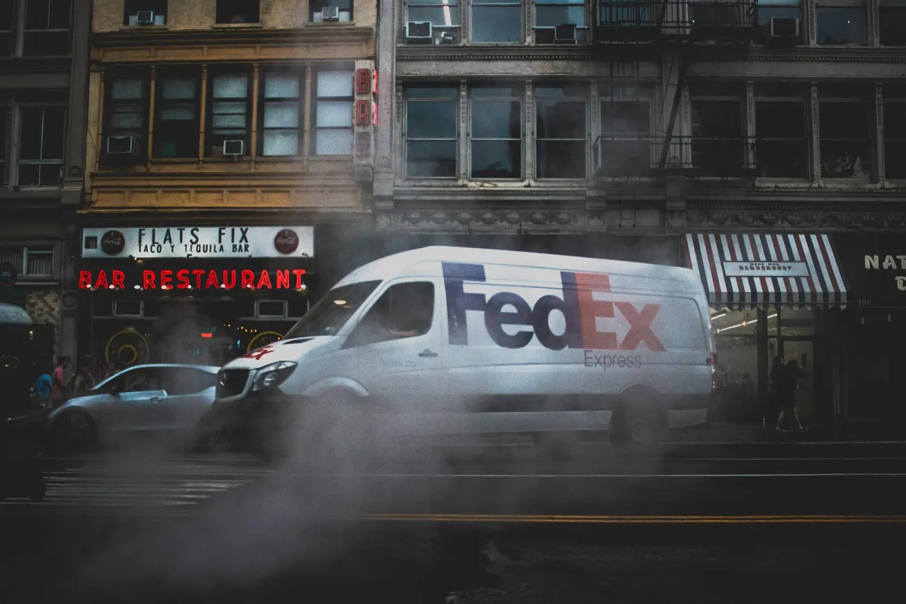 A FedEx van drives through a city street shrouded in mist, passing by urban buildings with vivid neon signs for a taco bar and restaurant. The scene feels moody and bustling.