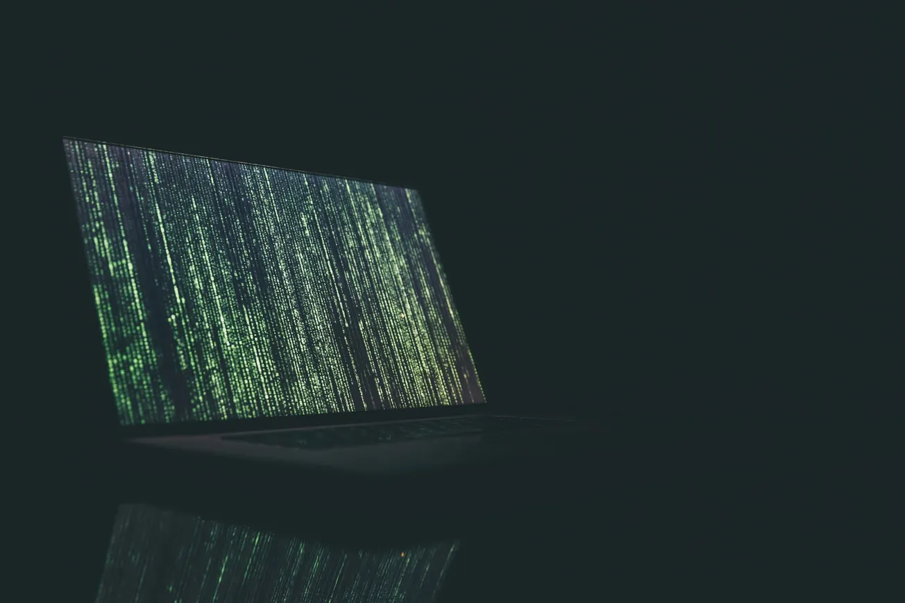 A laptop screen displays cascading green matrix-like code in a dark environment, conveying a mysterious and technological atmosphere.