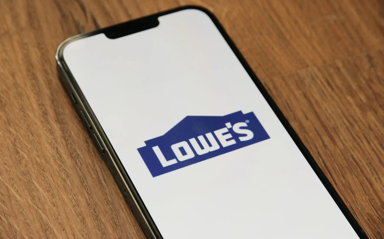 Smartphone displaying the Lowe's logo on its screen, placed on a wooden surface. The image conveys a professional and retail-focused tone.