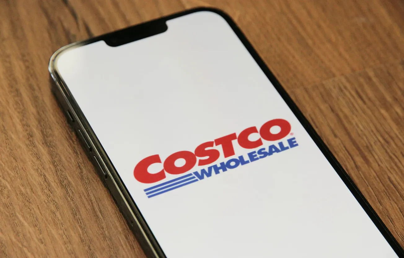 Smartphone displaying the Costco Wholesale logo on a bright screen, placed on a wooden surface. The mood is professional and business-oriented.