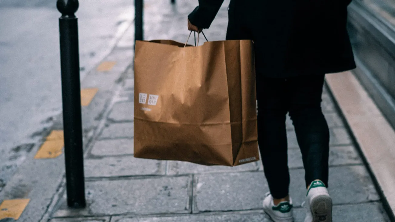 A person walks on a wet city sidewalk carrying a large brown paper shopping bag. The scene is casual and urban, with a cool, muted color palette.