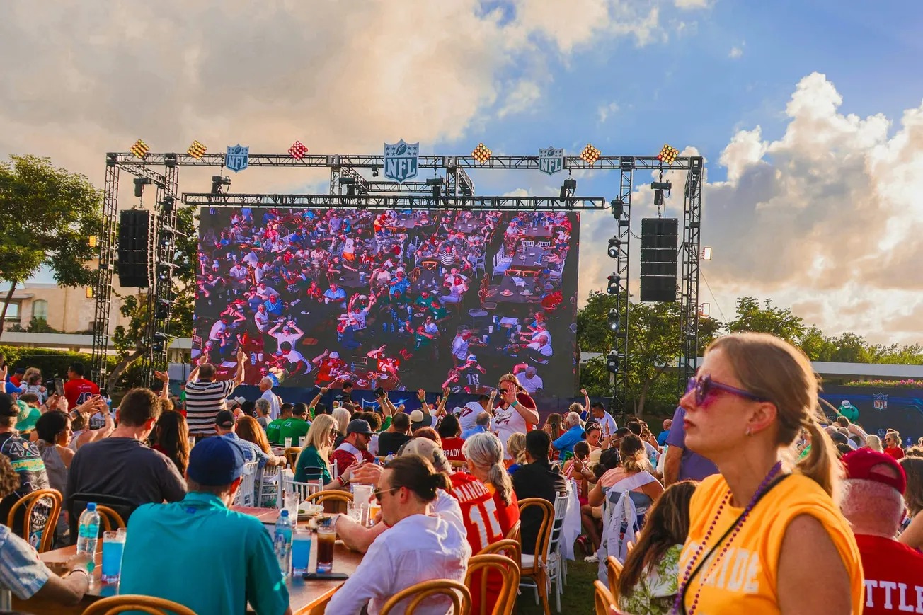 Crowd watching an outdoor NFL game on a large screen. People in jerseys sit at tables under a blue sky, creating a lively, festive atmosphere.