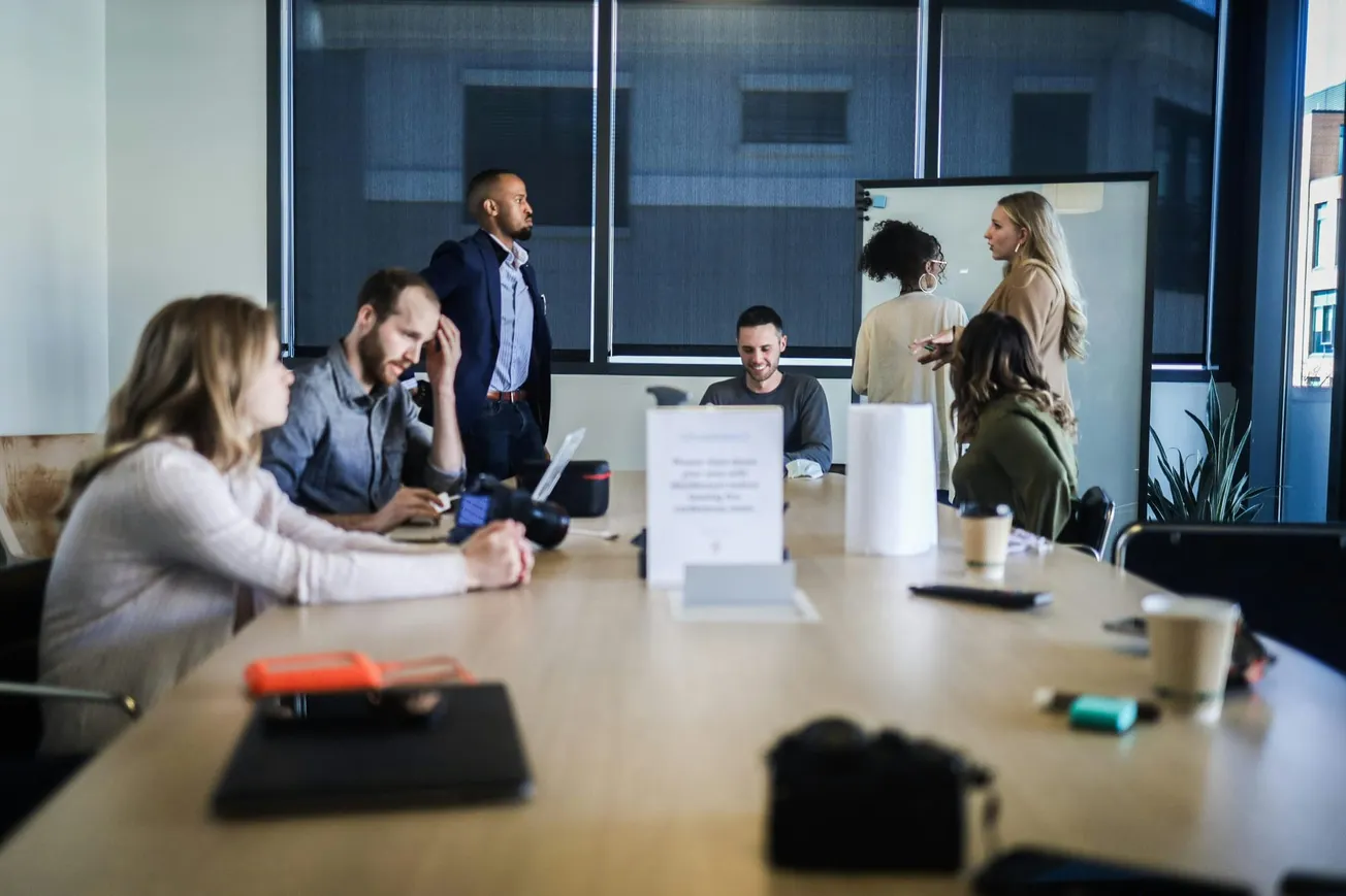 A diverse group of people in an office is having a meeting. Some are seated at a table with laptops, others are having a standing discussion by a whiteboard.