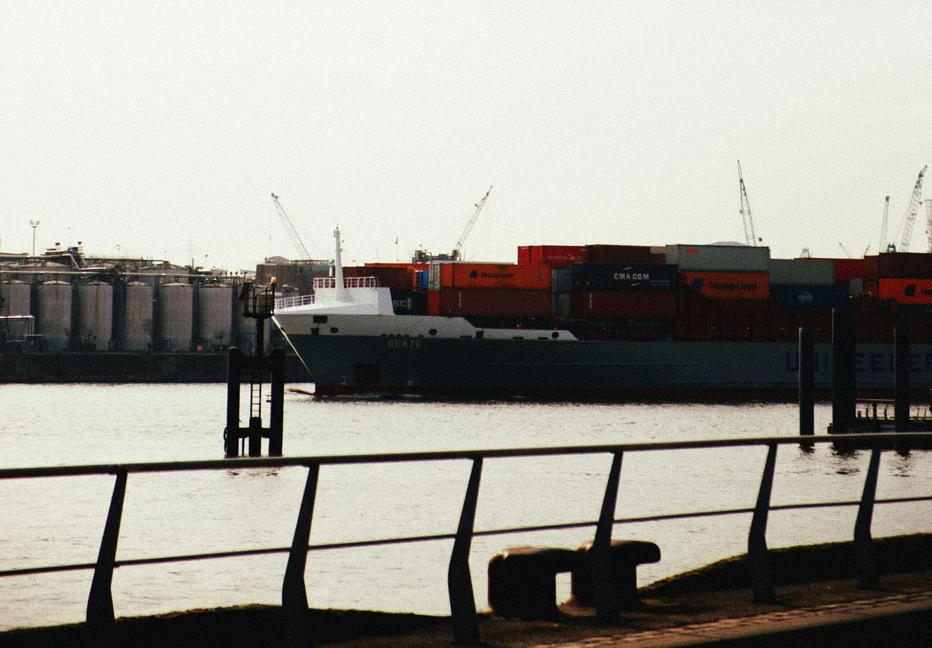 A cargo ship carrying colorful containers is docked in an industrial port. Silos and cranes are visible in the background under a cloudy sky. Calm, industrial scene.