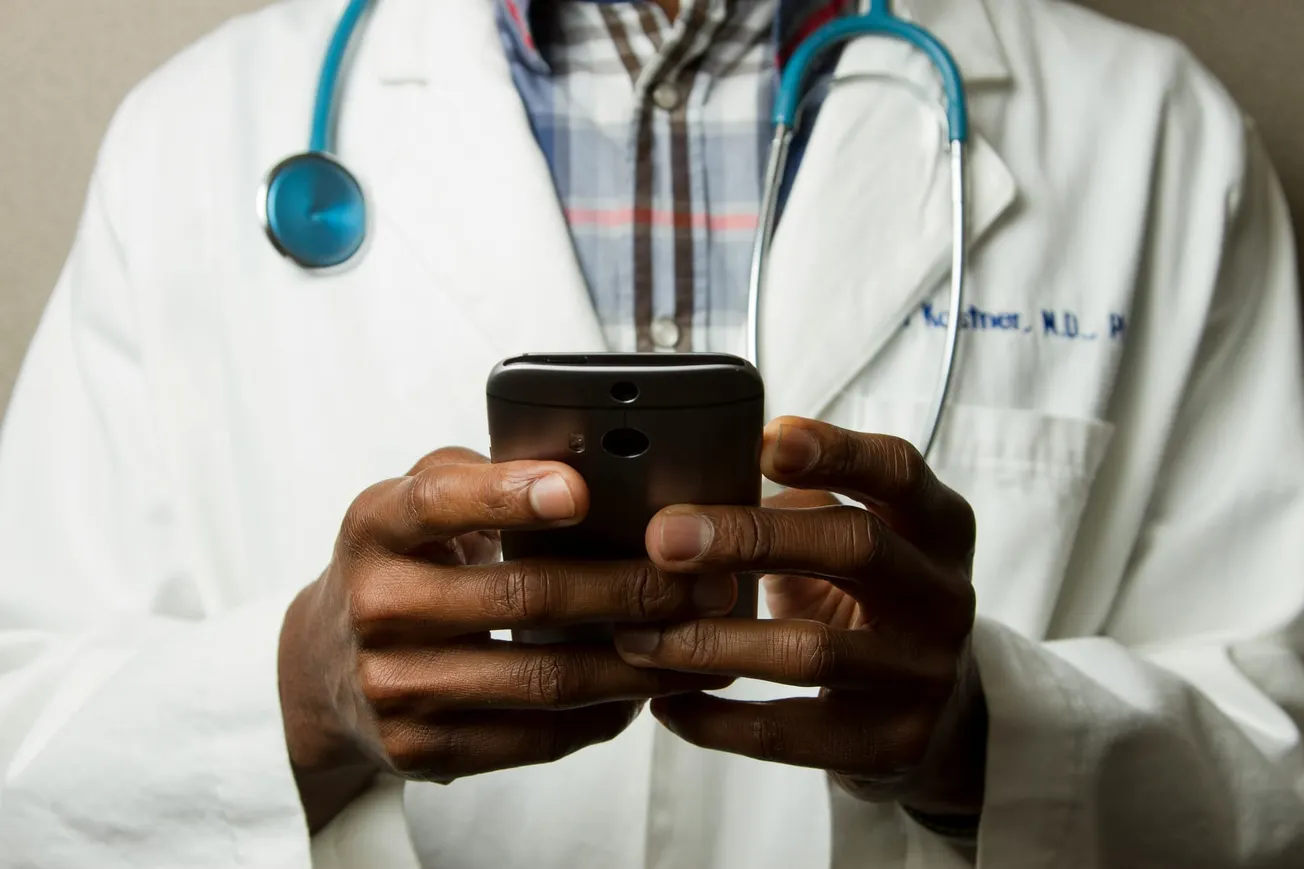 A doctor wearing a white coat and stethoscope holds a smartphone, focusing intently. The background is neutral, suggesting a professional setting.
