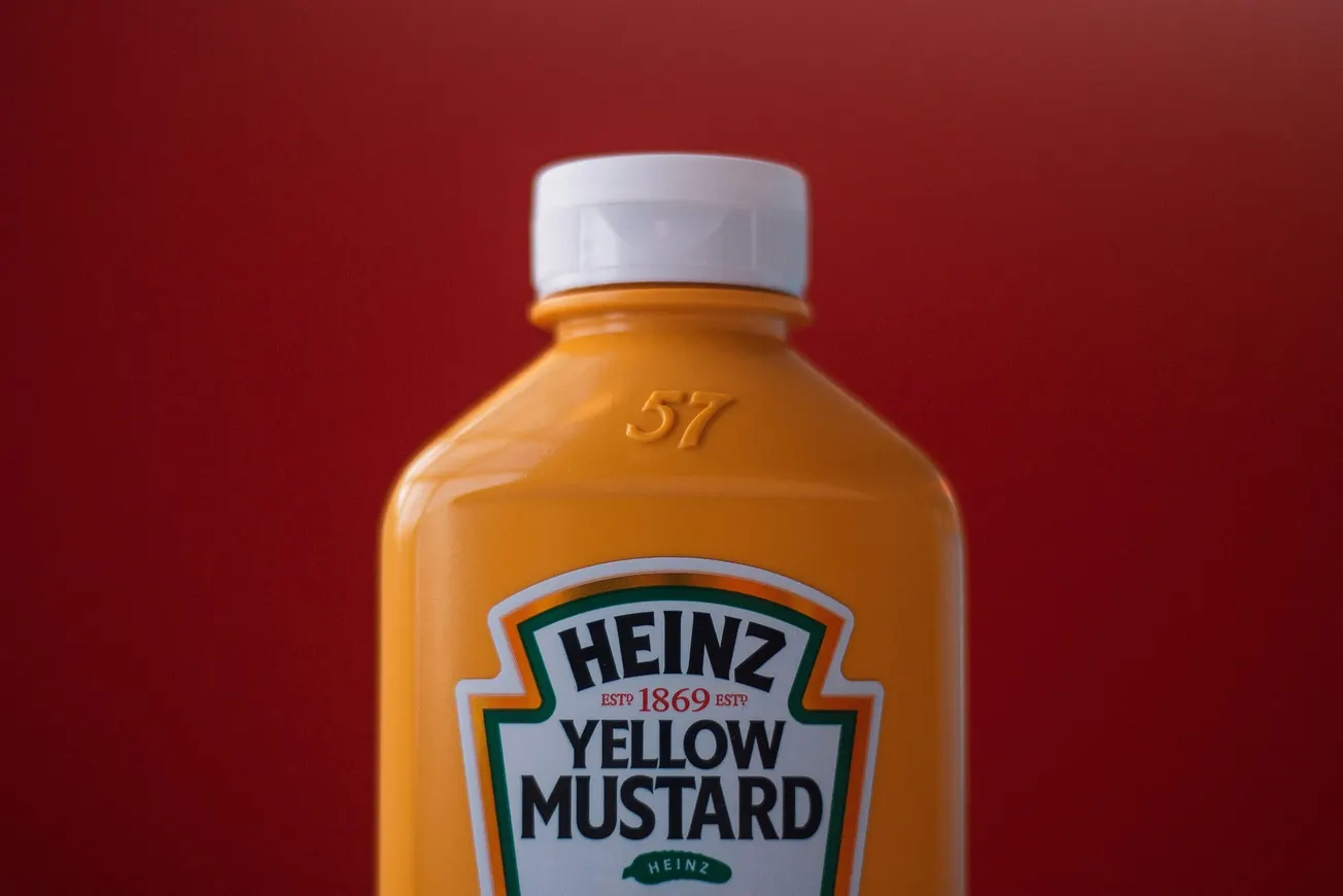 Bottle of Heinz Yellow Mustard with a white cap, shown against a deep red background. The mustard bottle is vibrant and eye-catching.