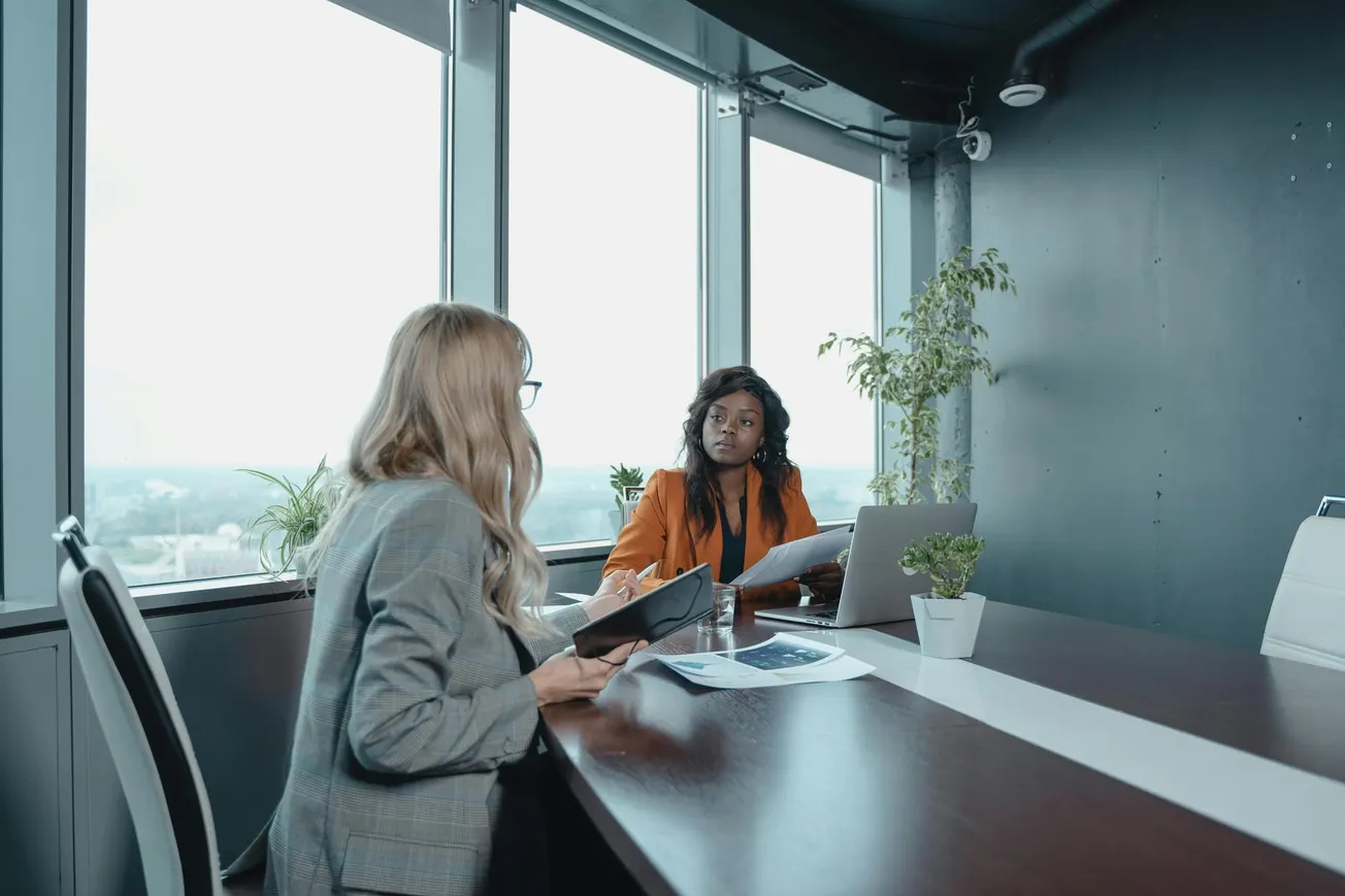 Two women in a modern office have a meeting. One, in a gray suit, takes notes, while the other, in an orange jacket, speaks. Bright windows behind them.