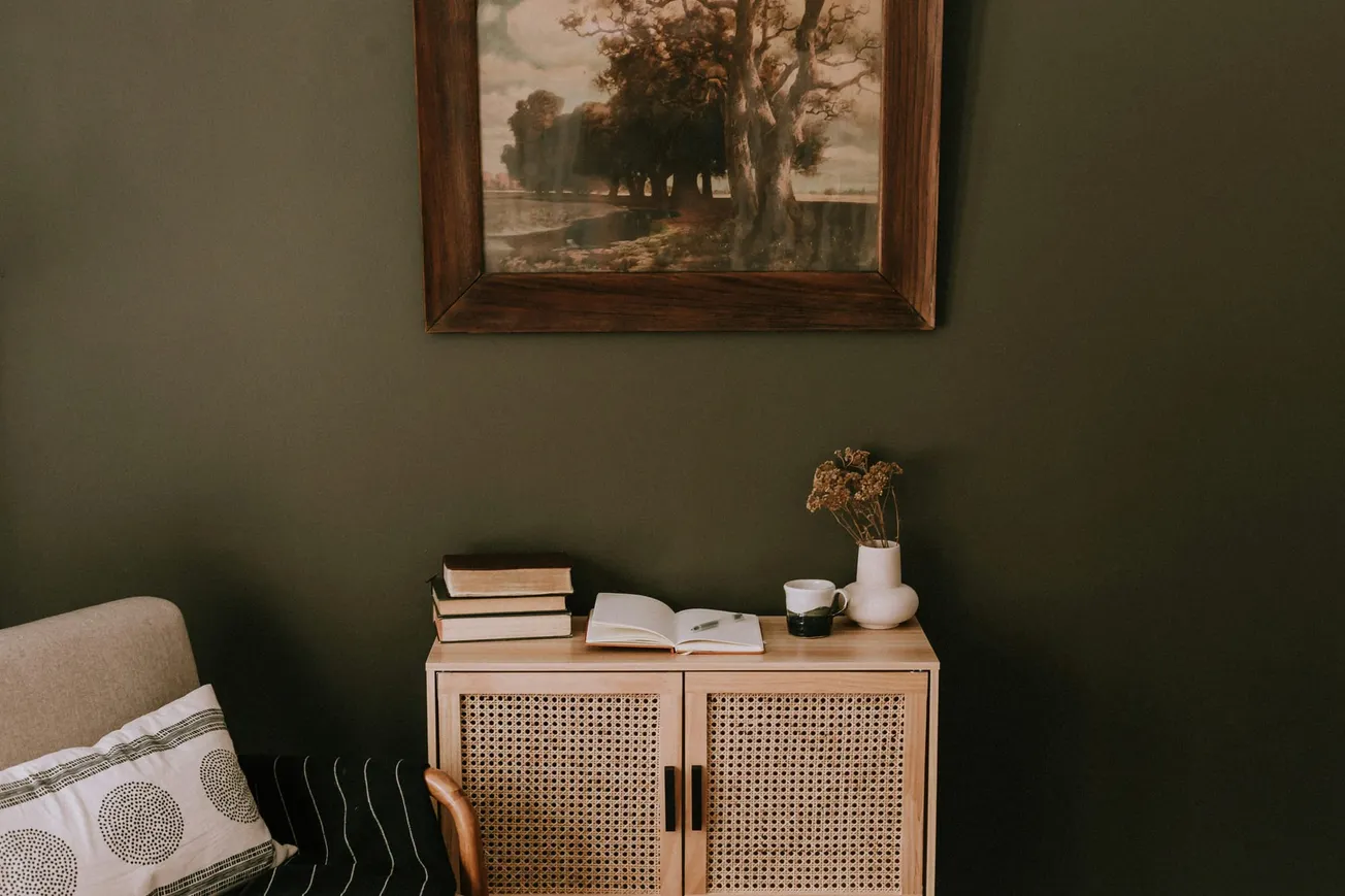 Cozy room with a wooden cabinet topped by books, a vase, and a cup. A vintage landscape painting hangs on an earthy green wall, creating a calm atmosphere.