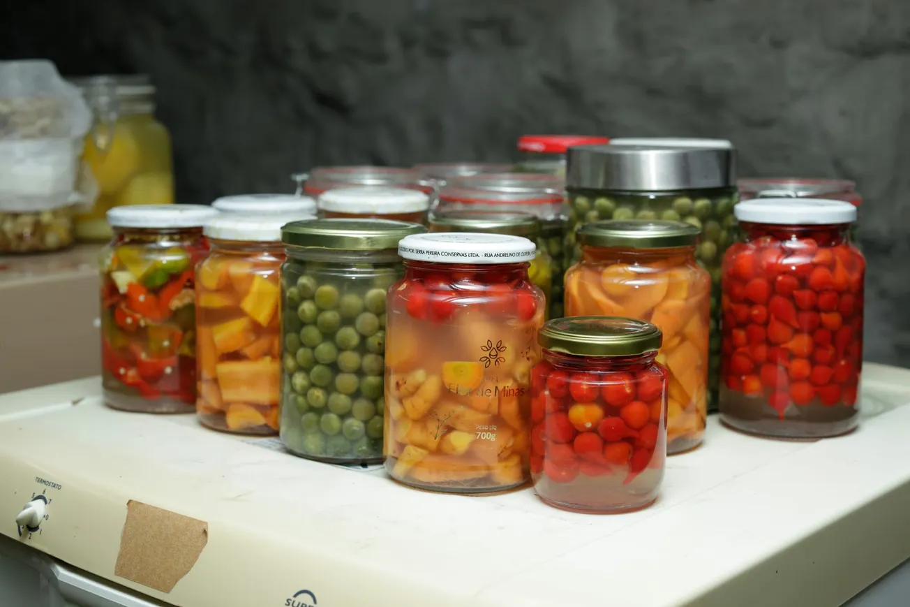 A collection of glass jars filled with preserved vegetables like peas, carrots, and peppers are arranged on a white surface against a gray stone background.