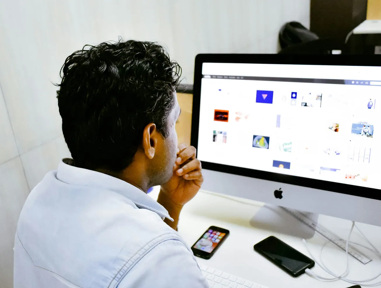 A man in a light blue shirt is focused on a computer screen displaying various images. Beside the computer are a smartphone and a tablet on the desk.