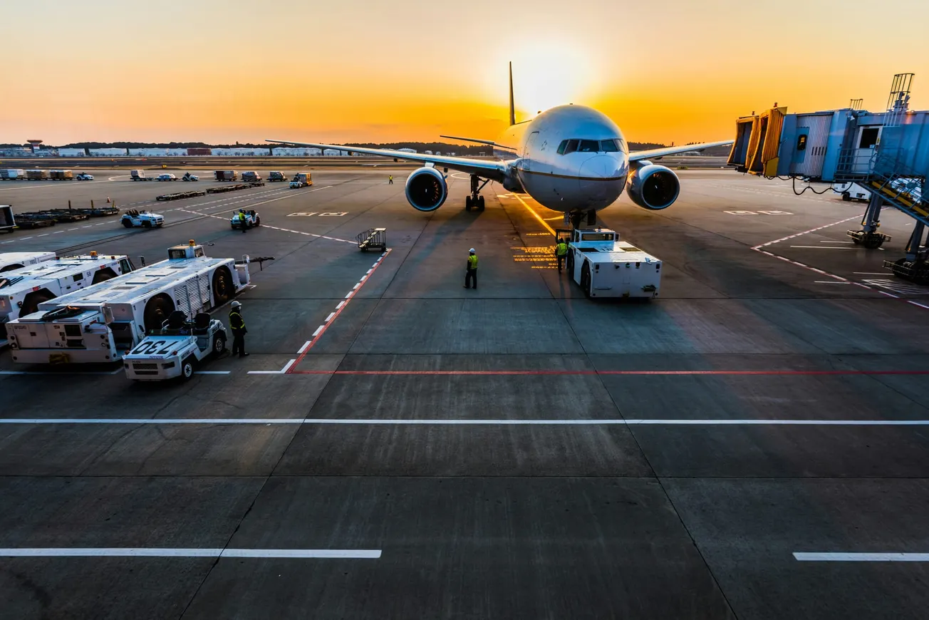 Airplane on tarmac at sunrise with vibrant orange sky. Ground crew and vehicles surround the aircraft near a terminal gate, creating a busy yet serene scene.