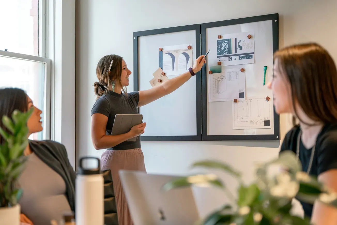 A woman gestures at charts on a bulletin board during a meeting, engaging colleagues seated around a table. The scene conveys collaboration and focus.