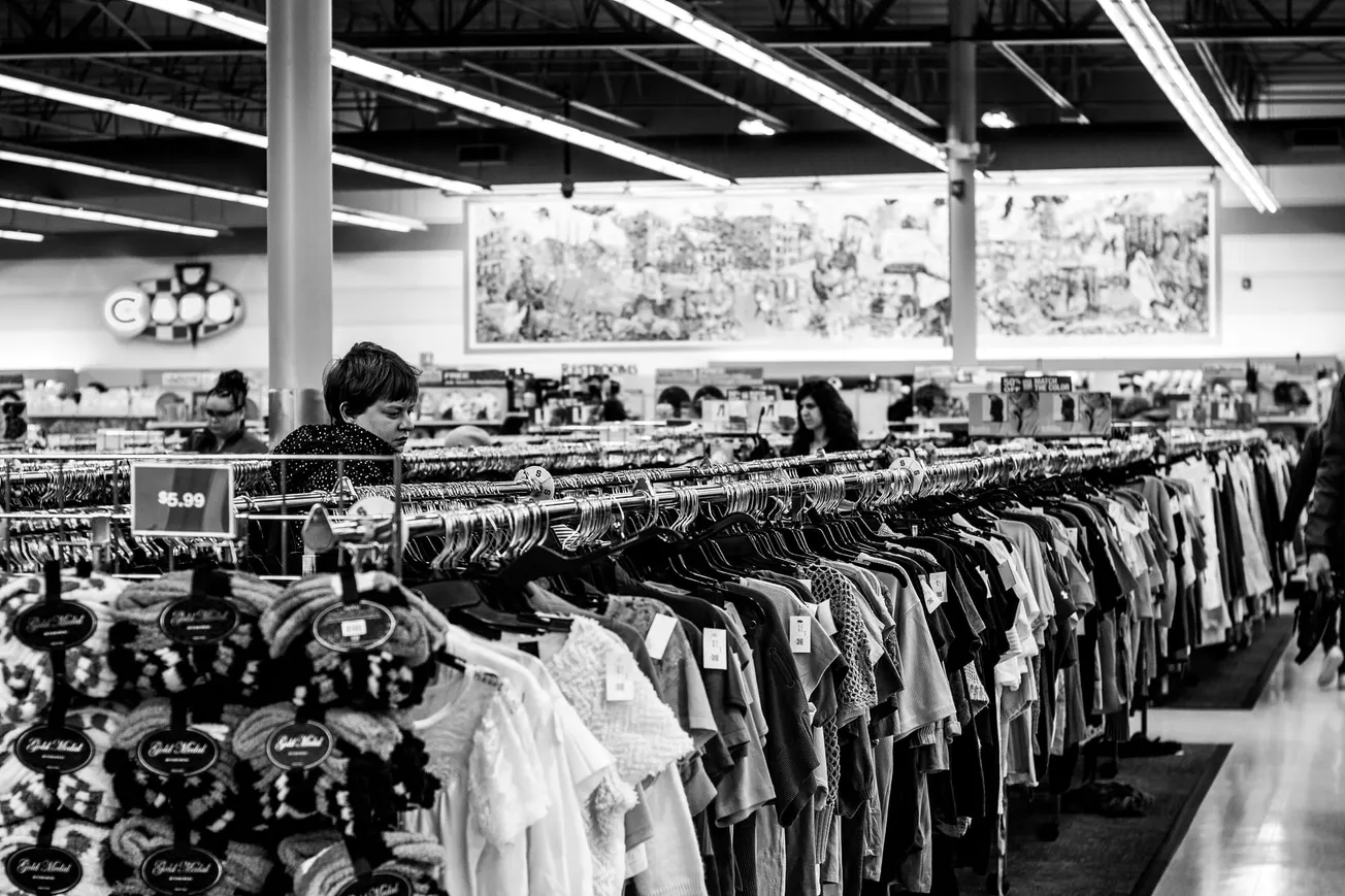 Black and white image of a thrift store with rows of hanging clothes. Shoppers browse racks under bright lights, creating a bustling, vintage ambiance.