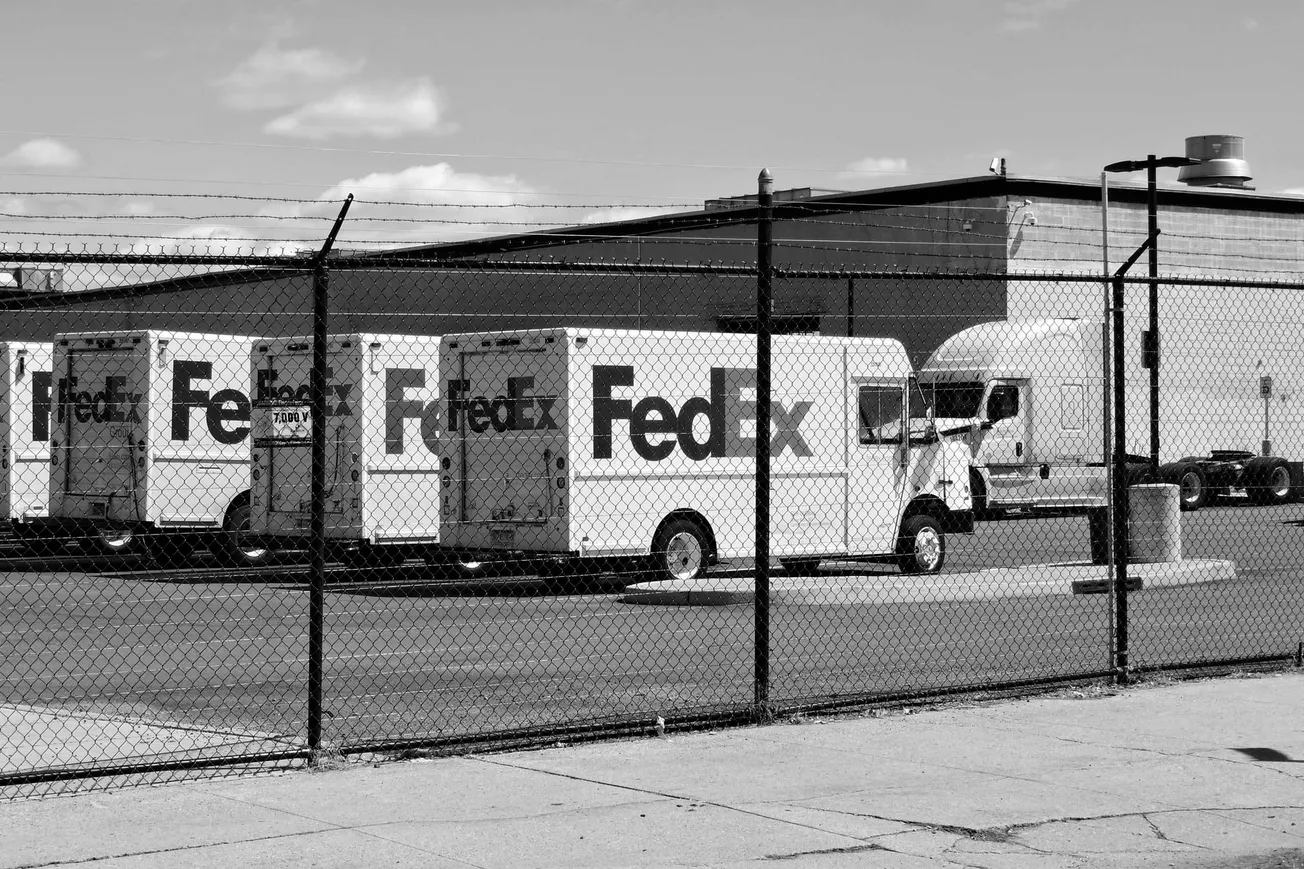 Black and white image of a fenced lot with multiple FedEx trucks parked in a row. An industrial building is visible in the background under a clear sky.