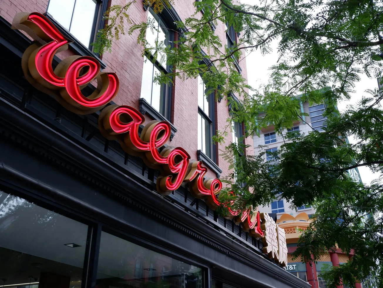 Red neon Walgreens sign on a historic building facade, surrounded by green leaves from nearby trees. The mood is lively and urban.