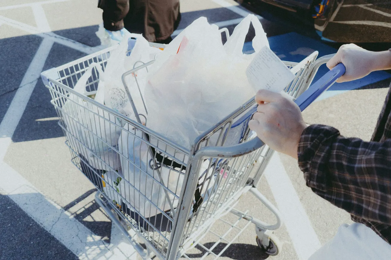 A person pushes a shopping cart filled with plastic bags in a parking lot. The person holds a receipt, suggesting recent purchases. Bright, sunny day.