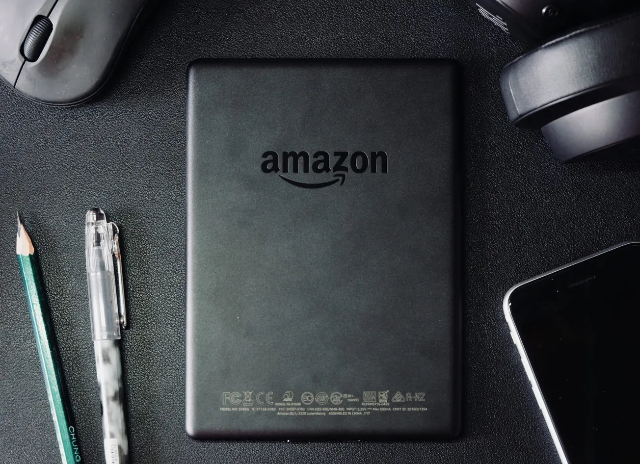 A black Amazon Kindle placed on a dark desk surrounded by a pencil, pen, computer mouse, headphones, and a smartphone, conveying a tech-focused mood.
