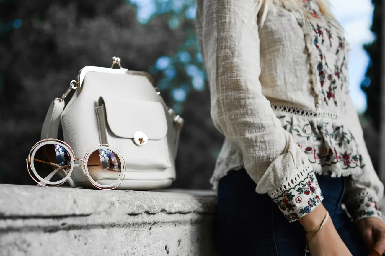 A stylish white bag and round sunglasses rest on a stone ledge next to a person in a patterned blouse, conveying a chic, casual vibe outdoors.