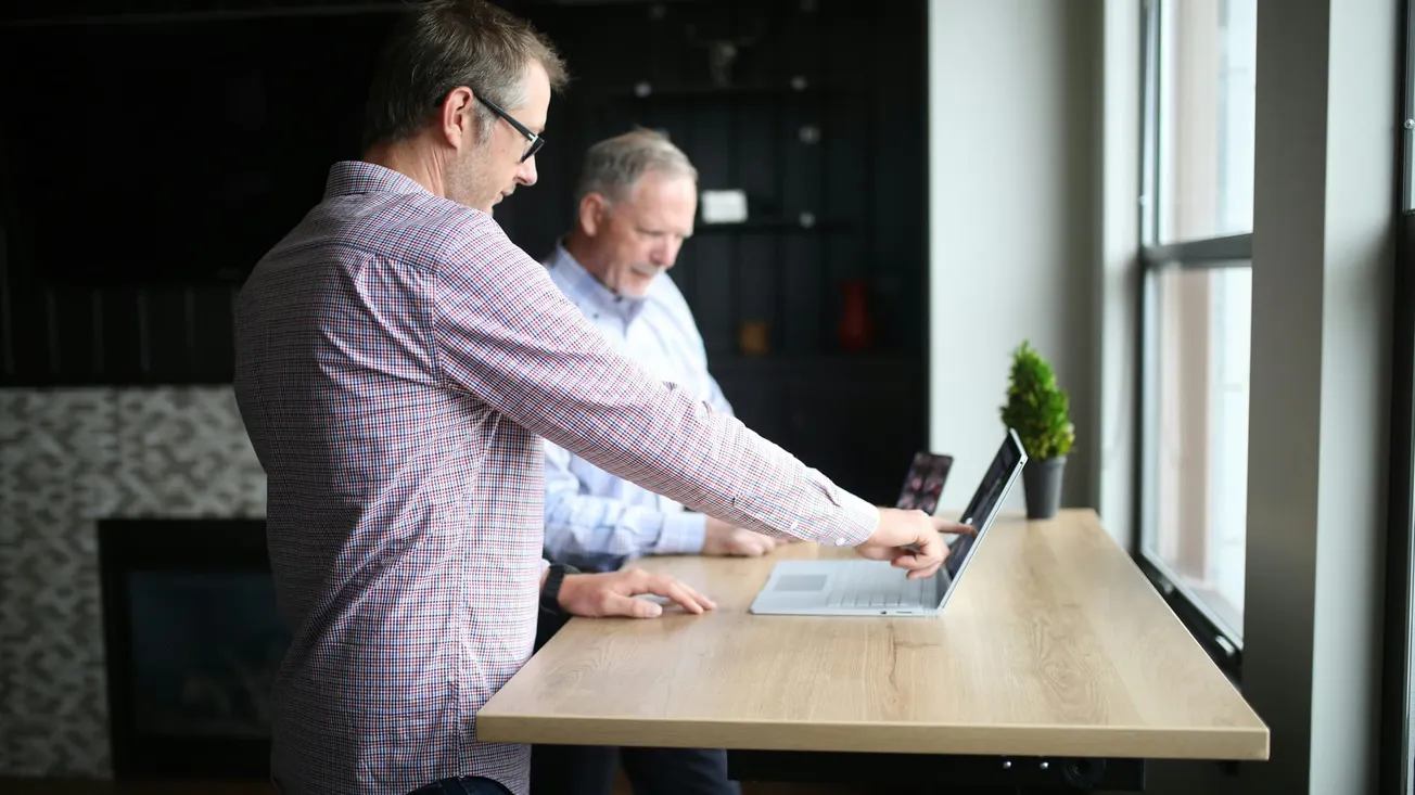 Two men stand at a wooden desk with a laptop, engaging in focused discussion. Sunlight streams through a window, creating a collaborative atmosphere.