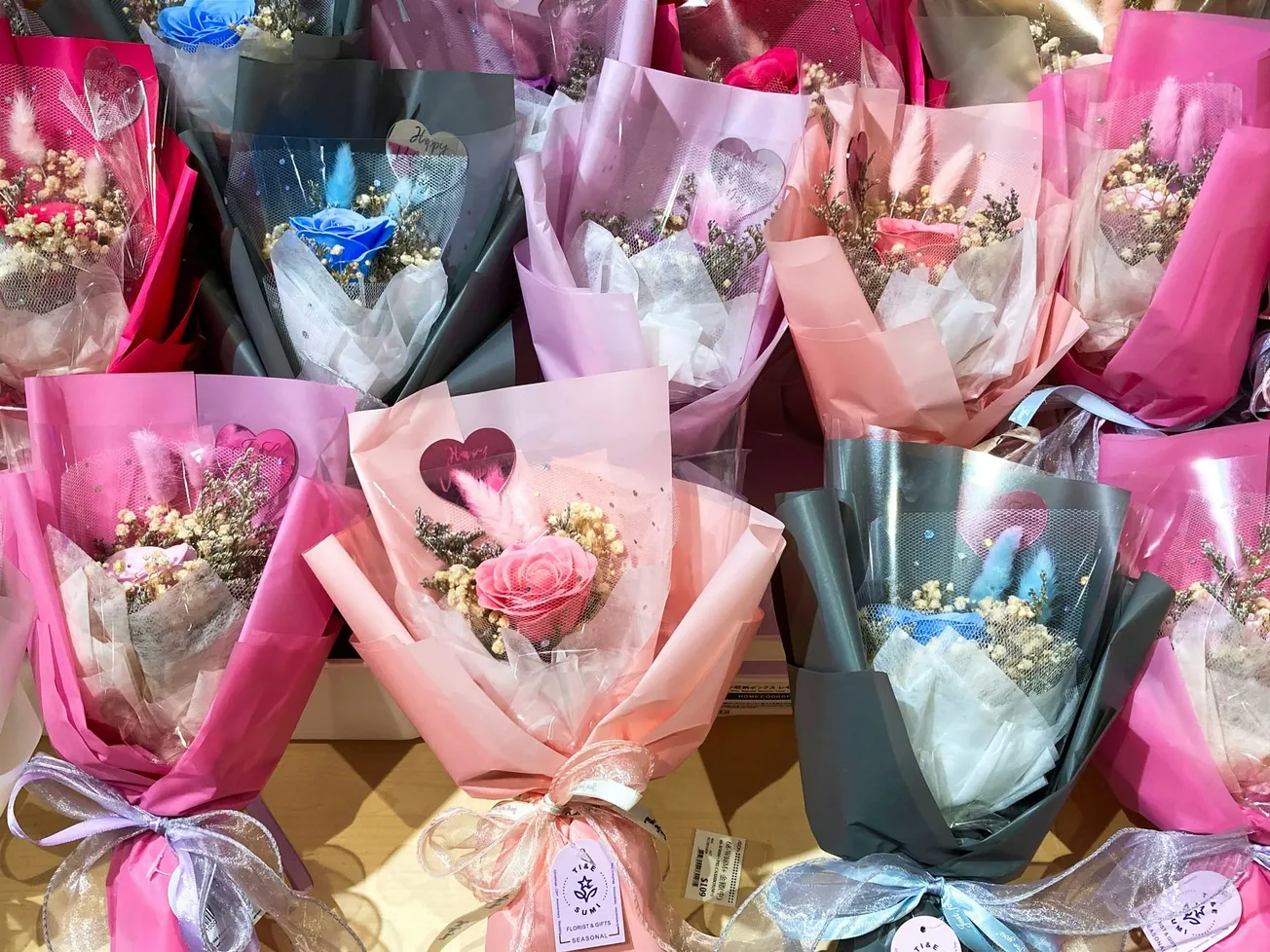 A variety of flower bouquets in pink, gray, and red wrapping with blue, pink, and red roses. They are adorned with baby's breath and ribbons.