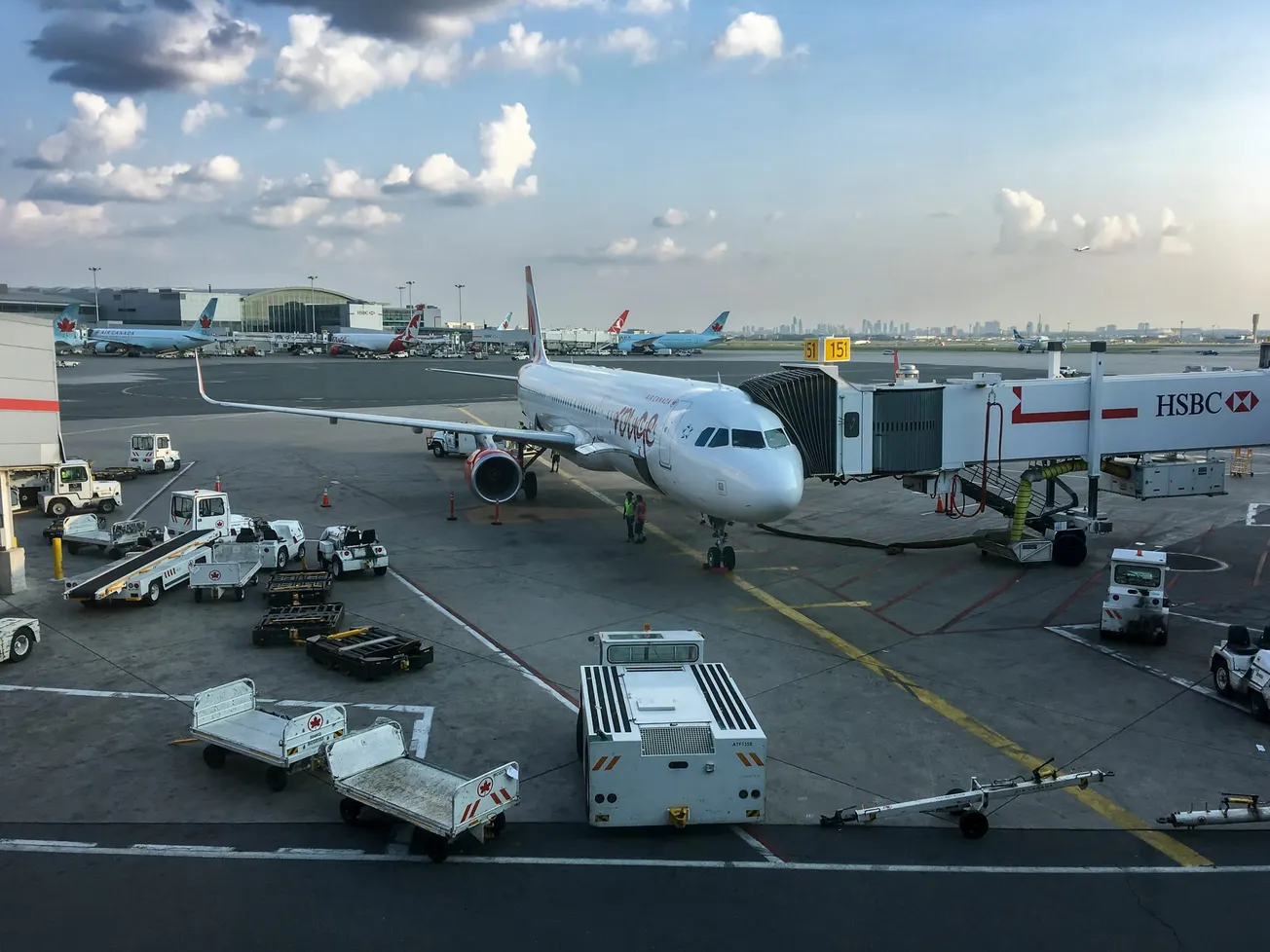 Airplane at airport gate with a jet bridge connected. Ground crew and vehicles are busy around the plane under a partly cloudy sky, creating a bustling atmosphere.