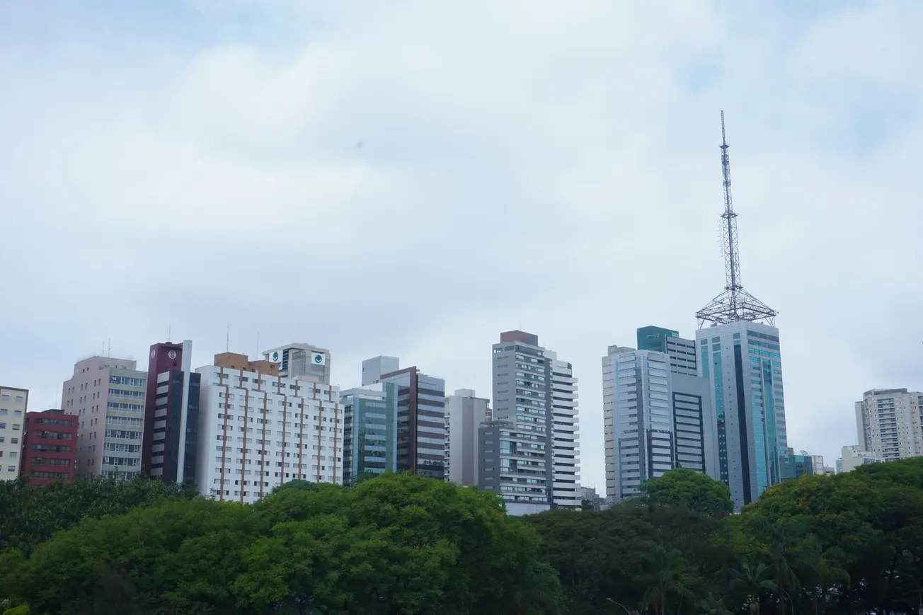 A city skyline featuring modern high-rise buildings under a cloudy sky. The foreground is lush with green trees, contrasting urban and natural elements.