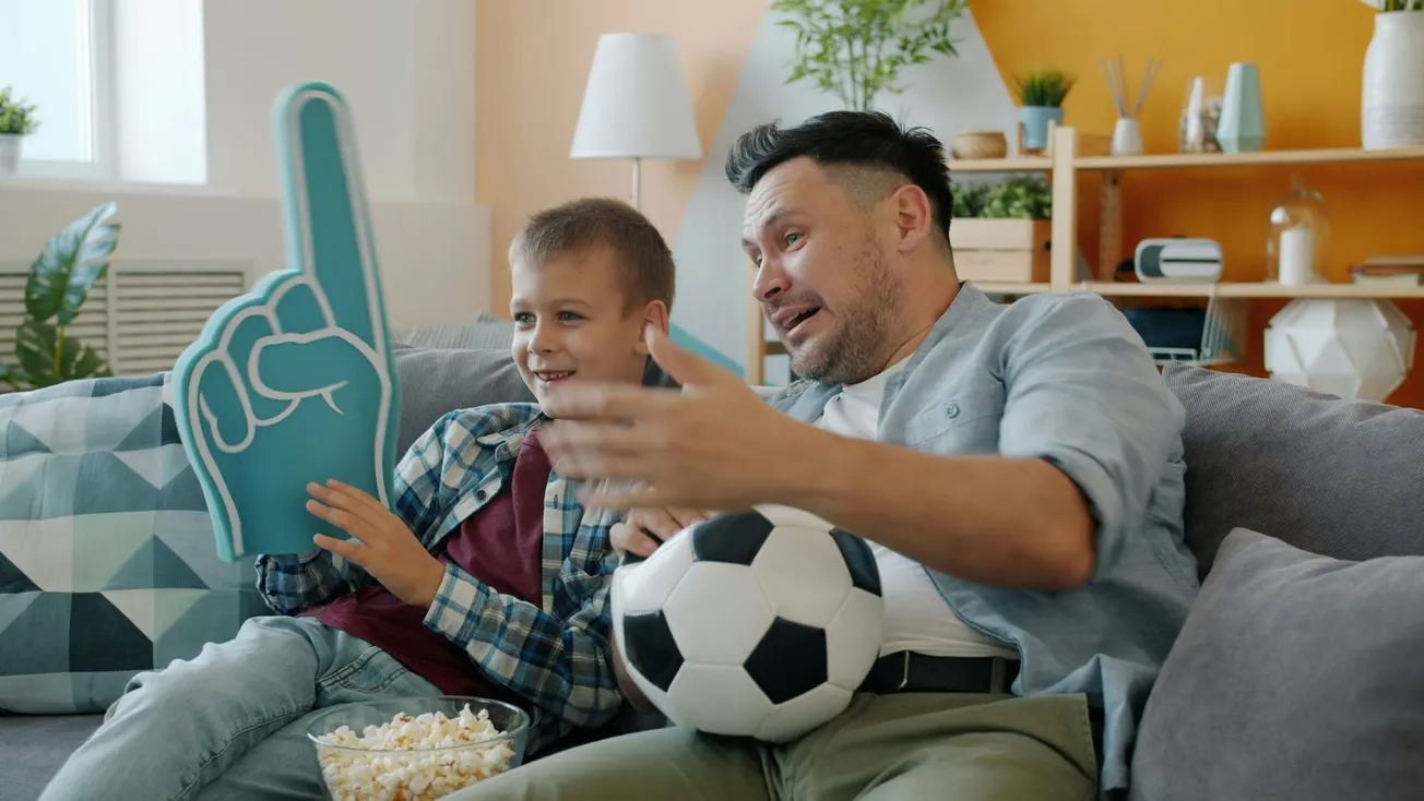 A man and a boy sit on a couch, enjoying TV. The boy holds a foam finger and popcorn, while the man holds a soccer ball, both looking excited.