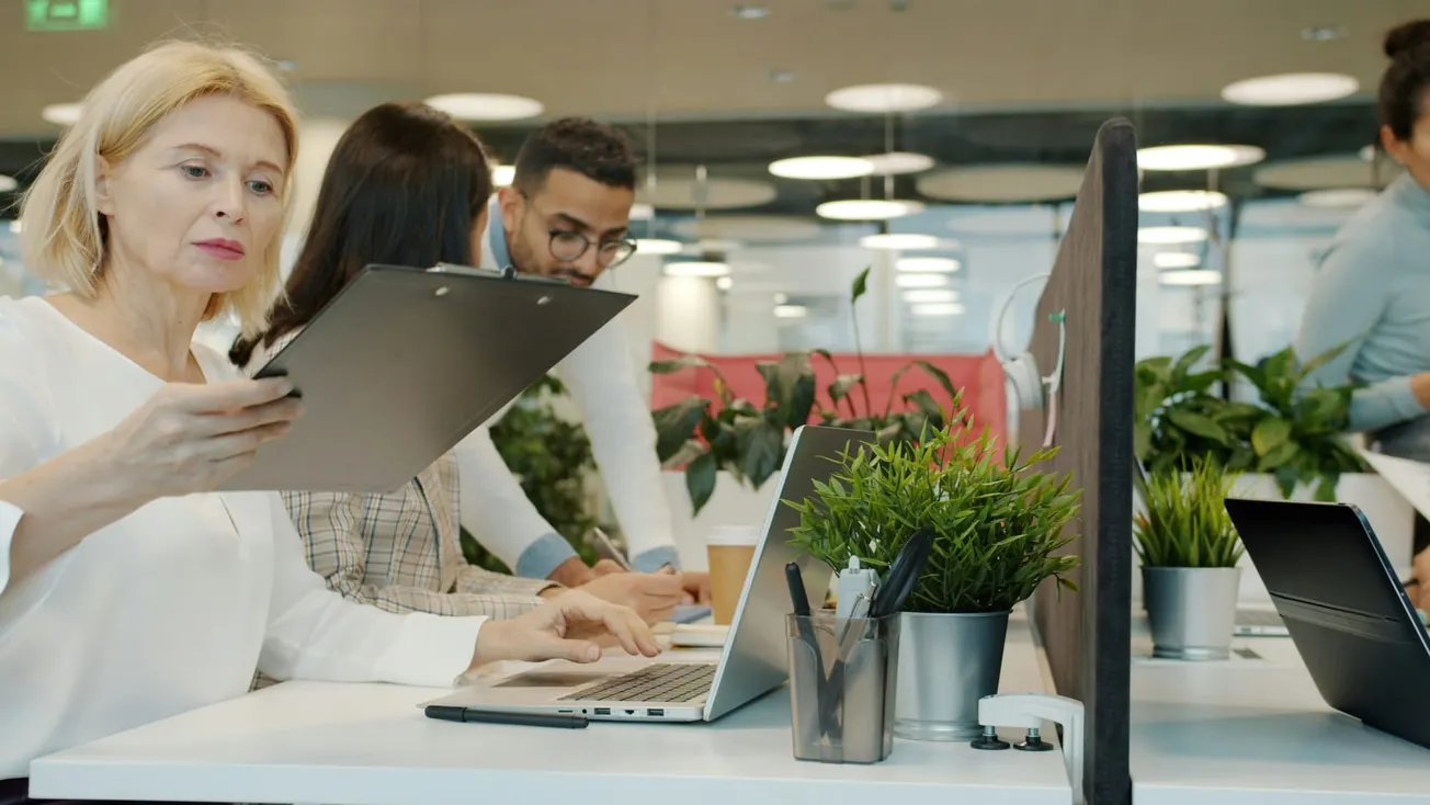 A diverse group of office workers in a modern setting. A woman examines a clipboard, others collaborate nearby. Laptops and plants create a focused yet lively atmosphere.