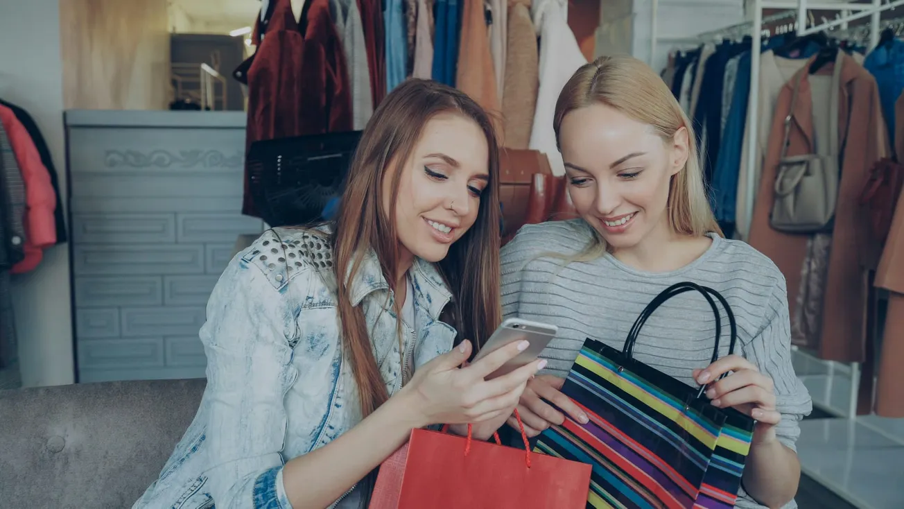 Two women sit smiling on a couch, holding shopping bags. One shows the other something on her phone. Clothing racks are visible in the background.