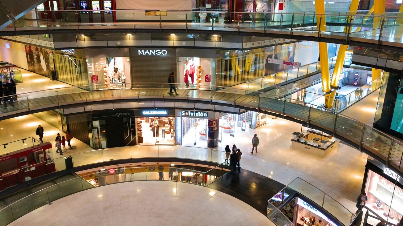A multi-level shopping mall with people walking past various lit store signs. The atmosphere is bustling and lively, with modern design elements.