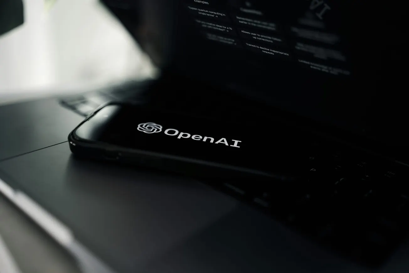 A smartphone displaying the OpenAI logo is placed on a laptop keyboard. The screen glows in dim light, suggesting a tech-focused, innovative atmosphere.
