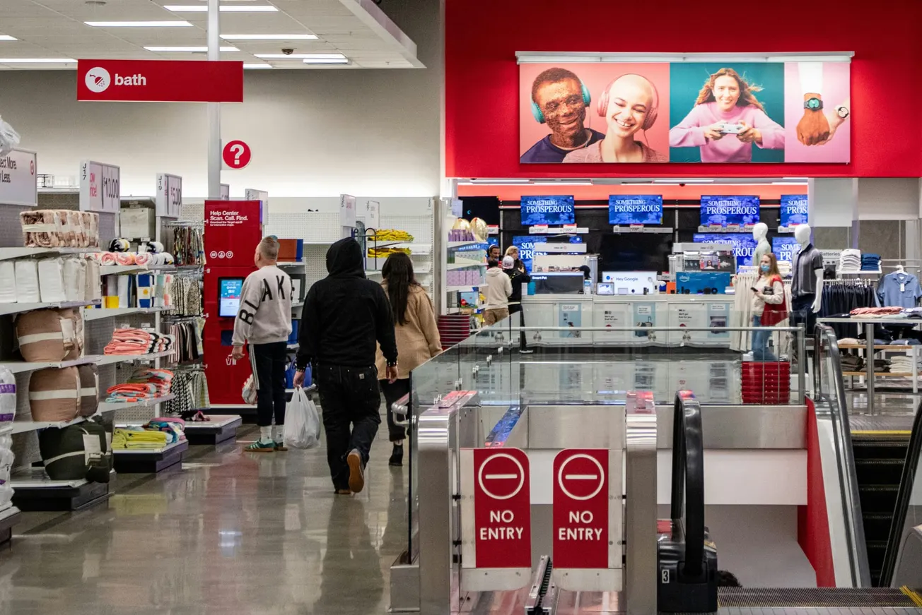 Shoppers in a brightly lit department store walk past bath goods. Large overhead ads feature smiling diverse faces. Escalator with "No Entry" signs in foreground.