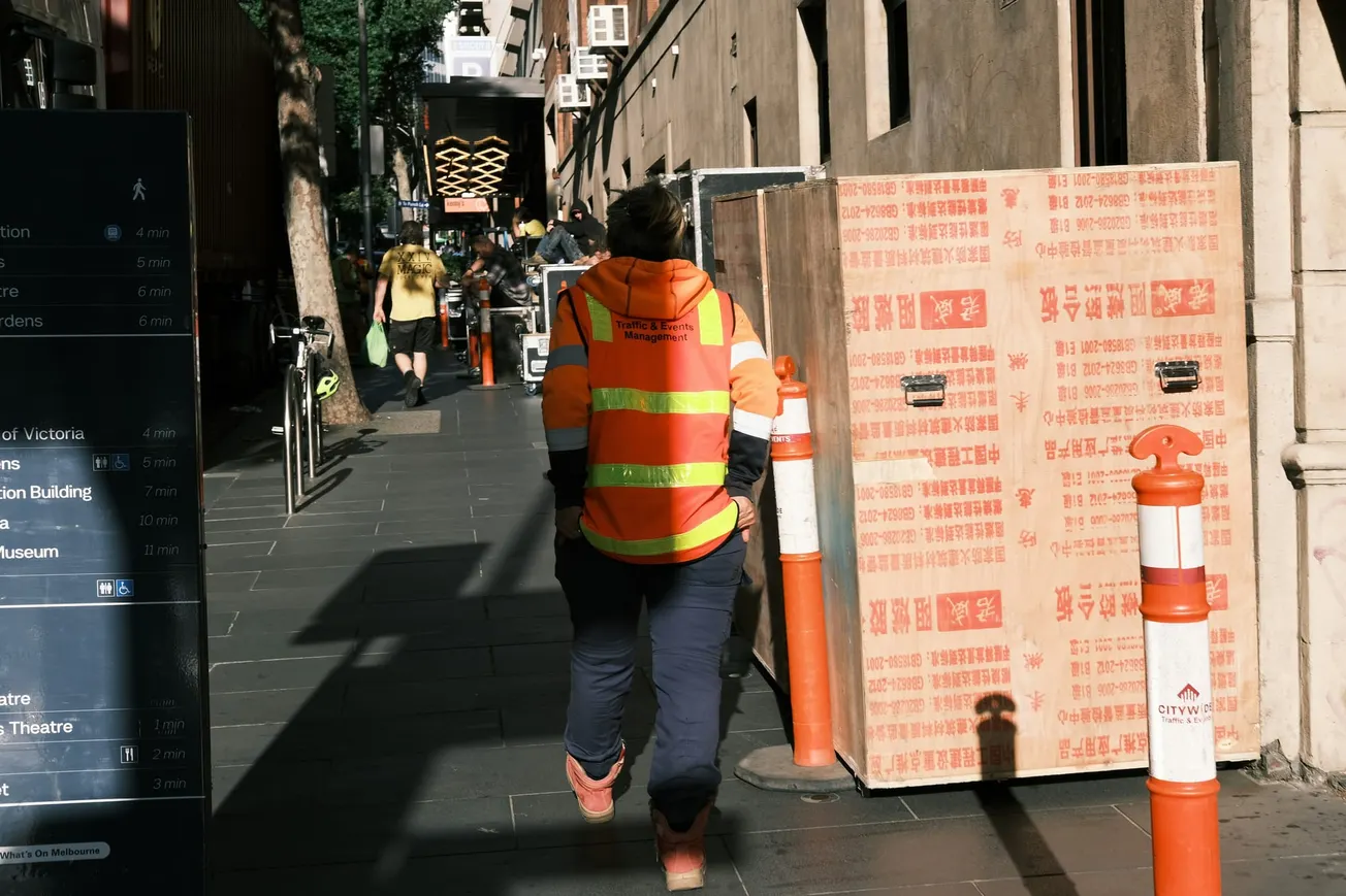 A person wearing a high-visibility orange safety vest labeled "Traffic & Events Management" walks down a city sidewalk past a large wooden crate and orange bollards.