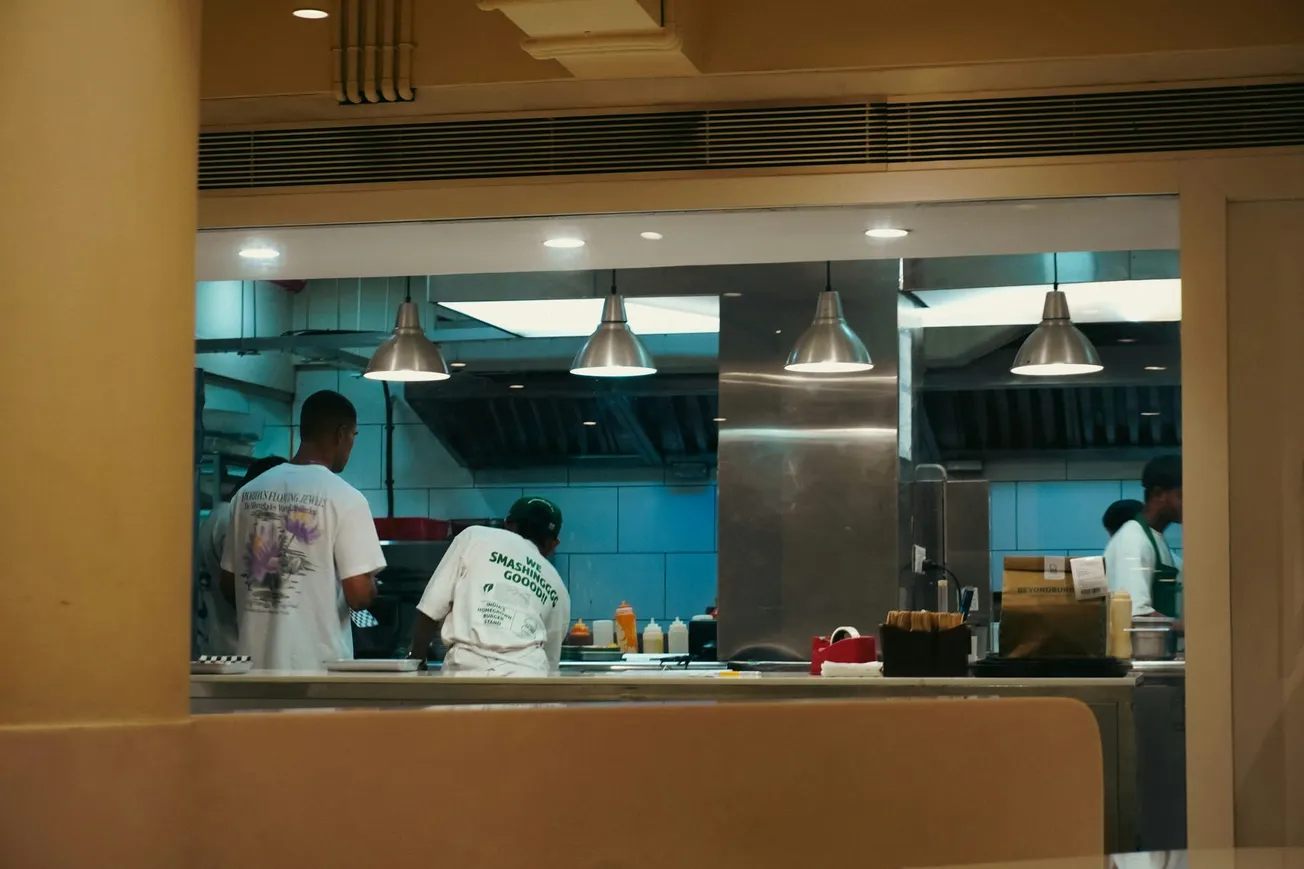 Restaurant kitchen with chefs in white shirts preparing food. Stainless steel surfaces, utensils, and condiment bottles are visible under bright overhead lights.