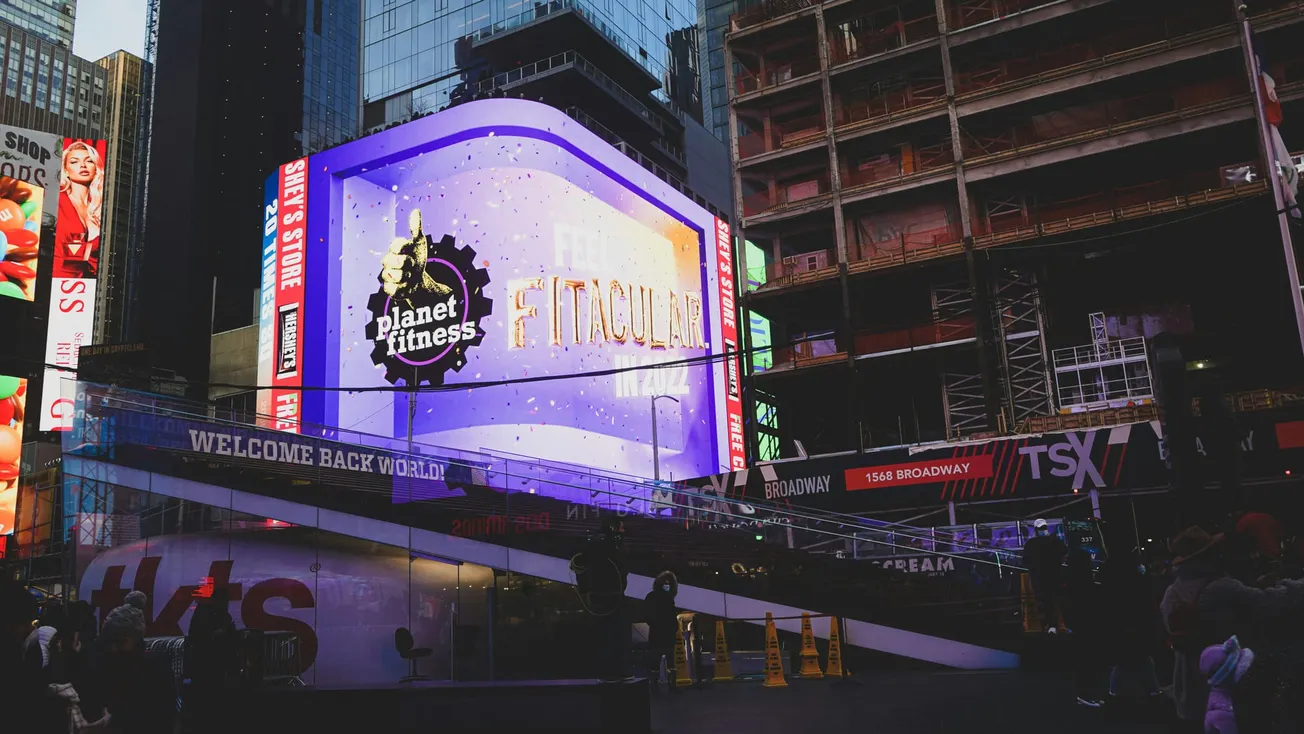 A large Planet Fitness billboard with "Fntacular" is lit up in a bustling urban setting. Tall buildings and colorful ads create a lively atmosphere.