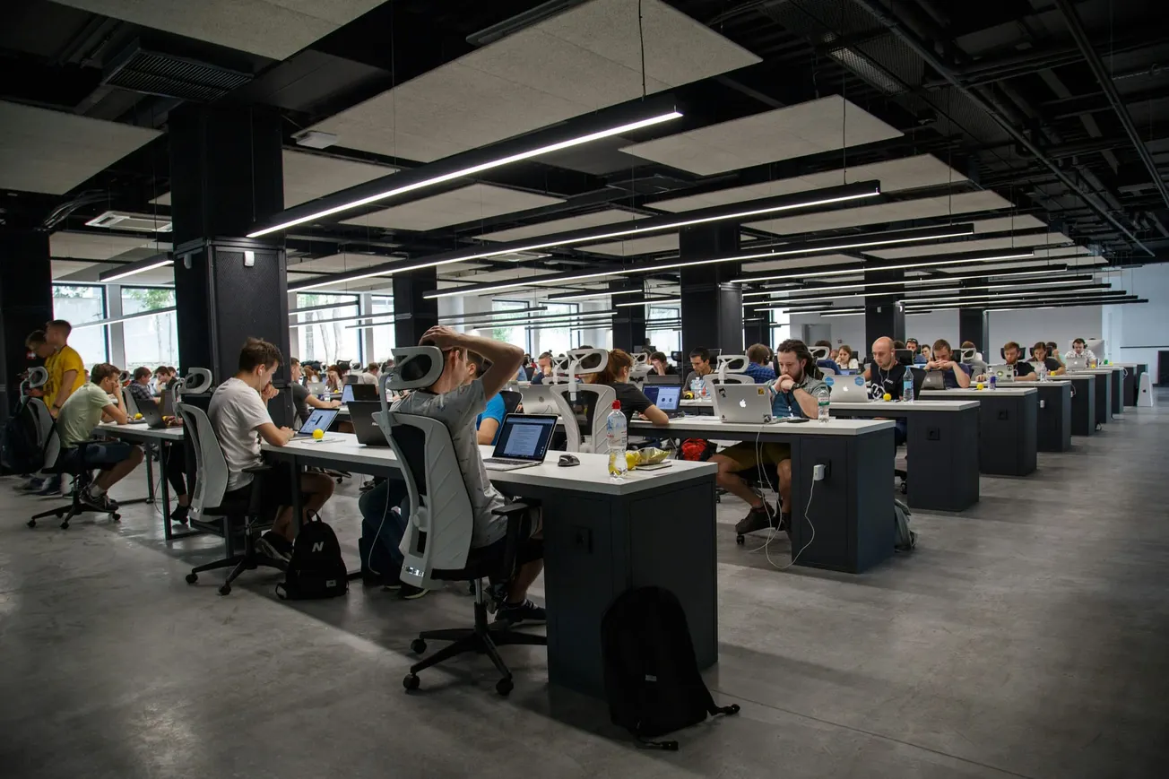 Large open office with many people working at rows of desks on computers, under fluorescent lights, in a modern workspace environment.