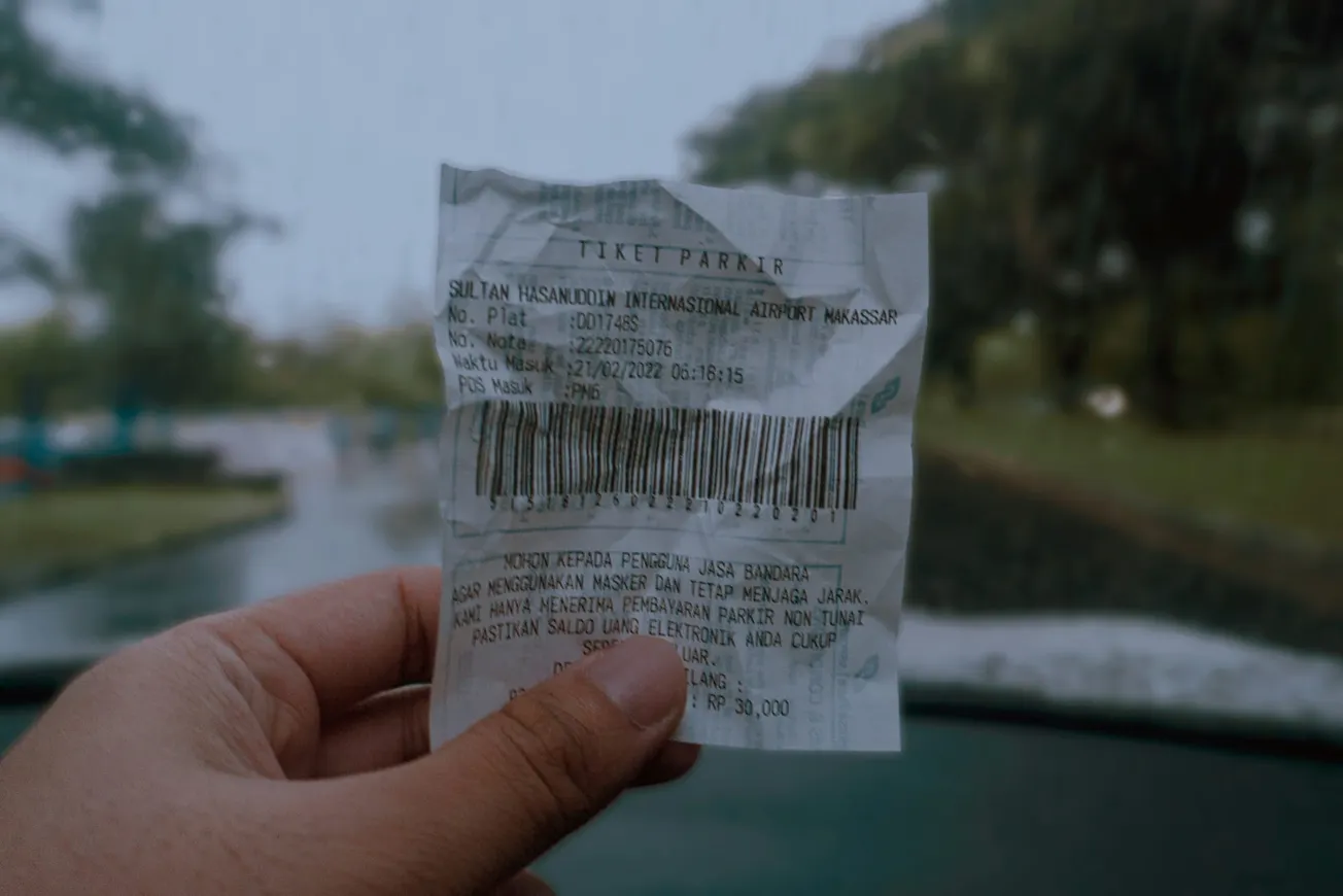 A hand holds a parking ticket for Sultan Hasanuddin International Airport. Blurry, rainy background of a road seen through a windshield. Moody atmosphere.