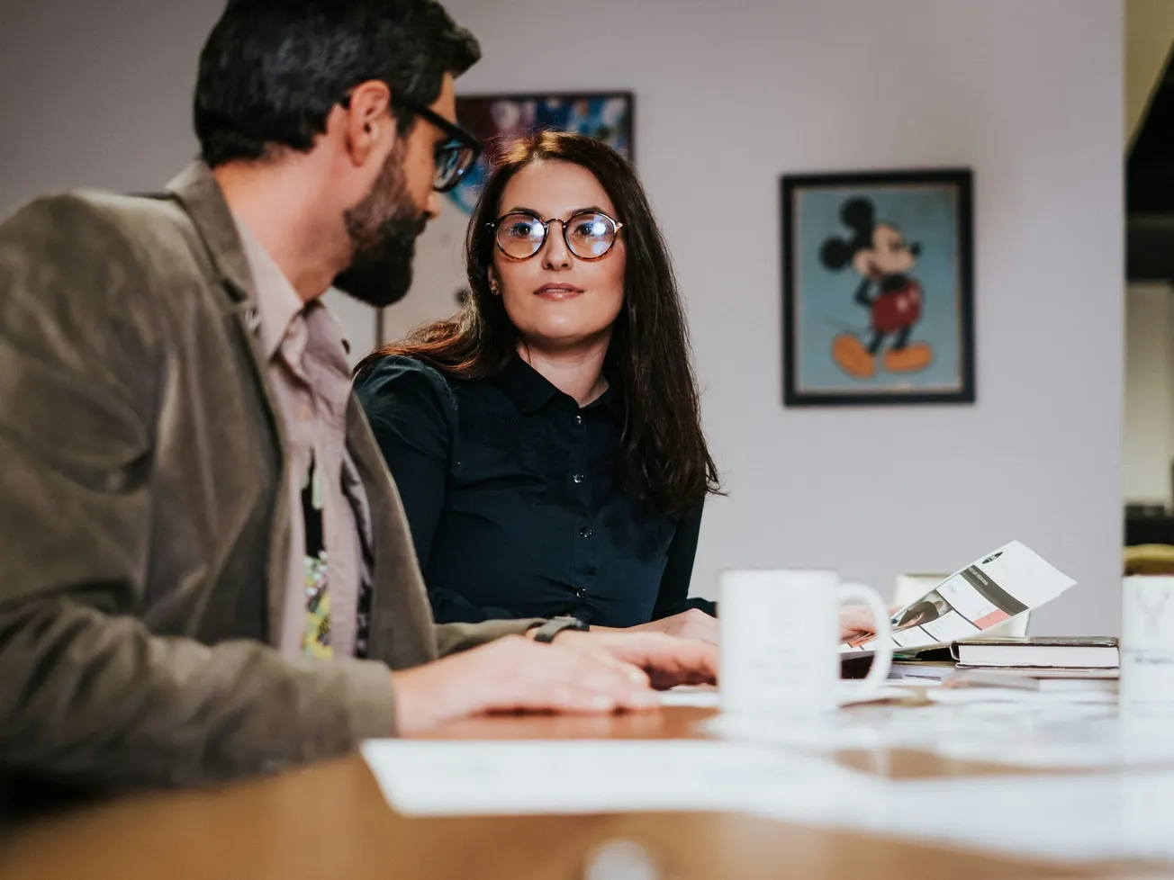 Two colleagues in an office, engaged in discussion. A woman in glasses looks attentively at a man. A Mickey Mouse poster is on the wall.
