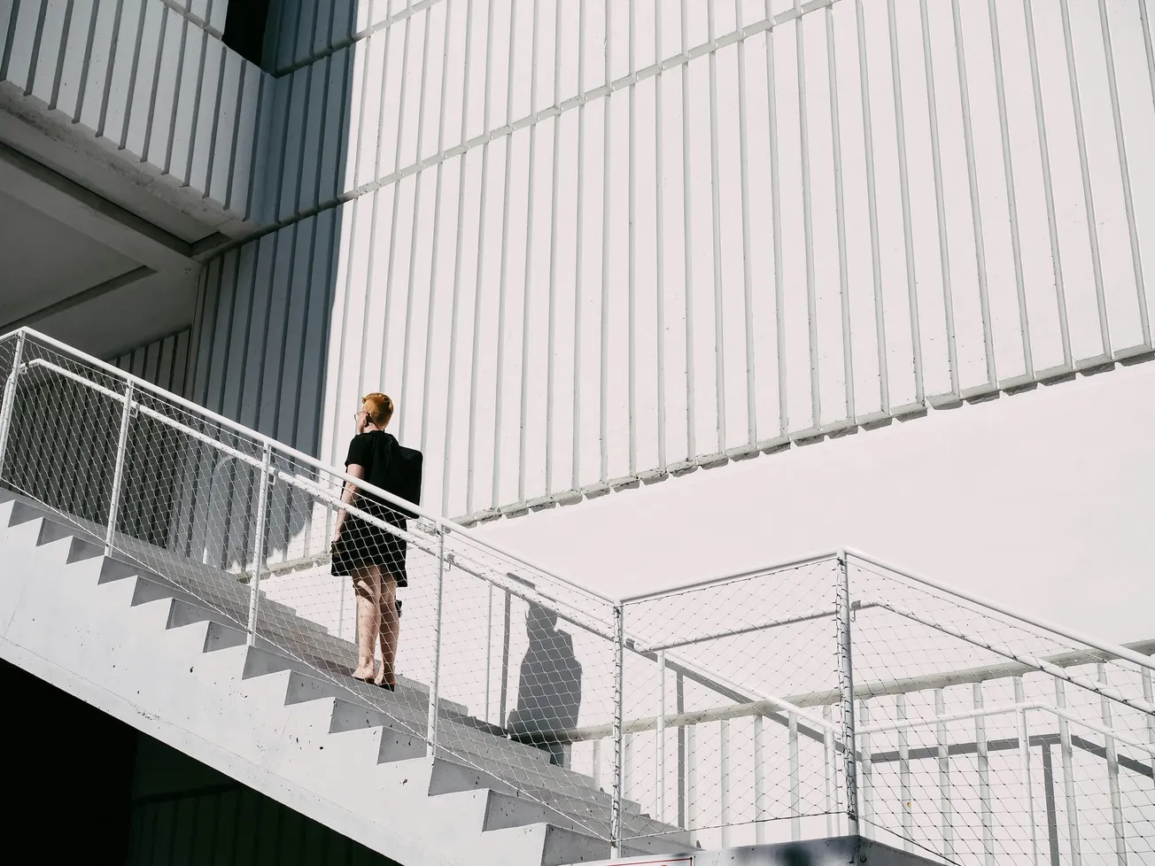 A person in a black dress ascends a sunlit, external staircase against a white, textured building. The scene feels minimalistic and modern.